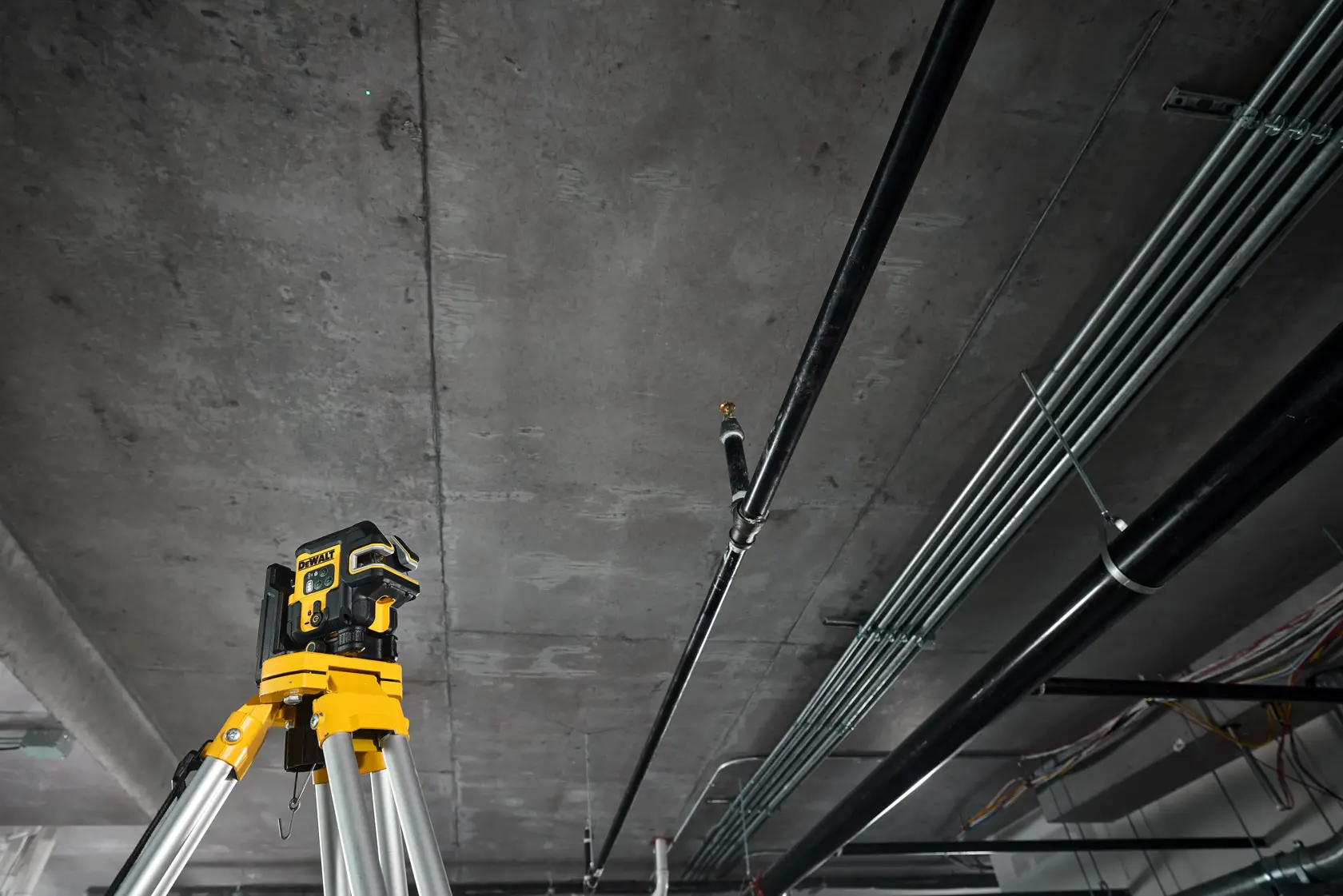 A DEWALT laser level device mounted on a tripod is positioned in an industrial or construction environment, pointing toward a ceiling with various exposed pipes and cables.