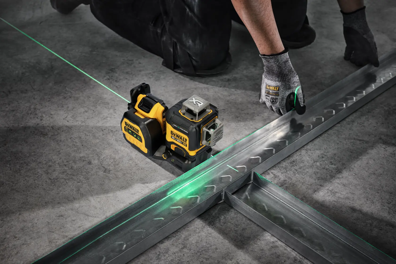 A DeWalt laser level device projects a green laser line onto a concrete floor, assisting a worker wearing gloves to align metal framing pieces.