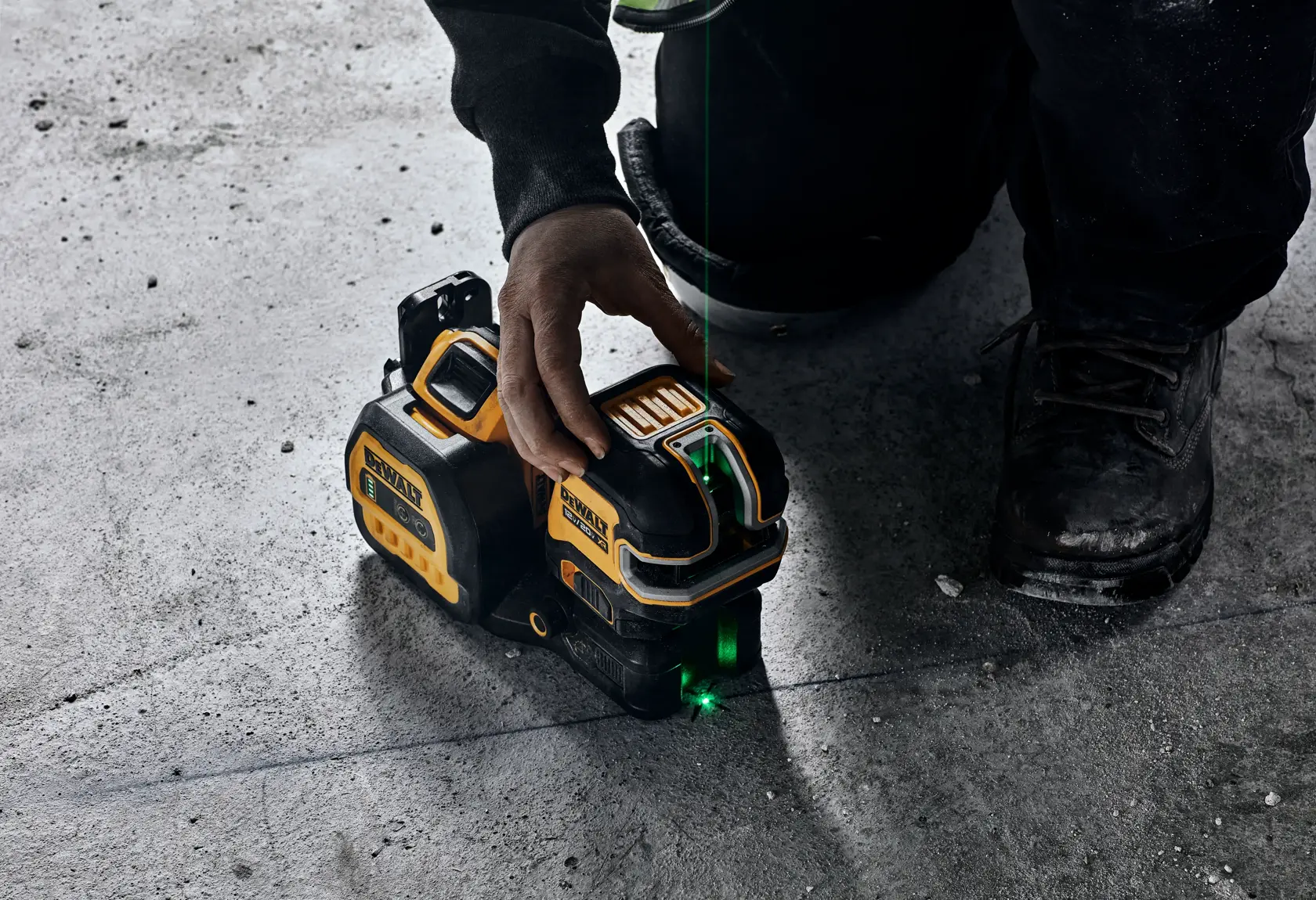 A close-up of a person using a DEWALT laser level on a concrete floor. The device emits a green laser line for alignment, and the person is operating it by hand while kneeling.