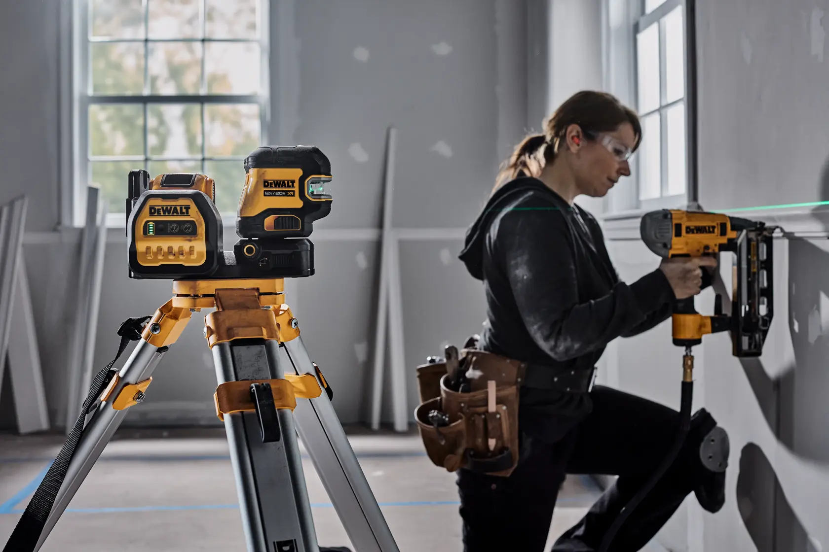 A person with a tool belt is using a DEWALT nail gun to work on a wall near a window, while a DEWALT laser level mounted on a tripod projects a green line across the wall for alignment. The scene appears to be in a room under construction or renovation.