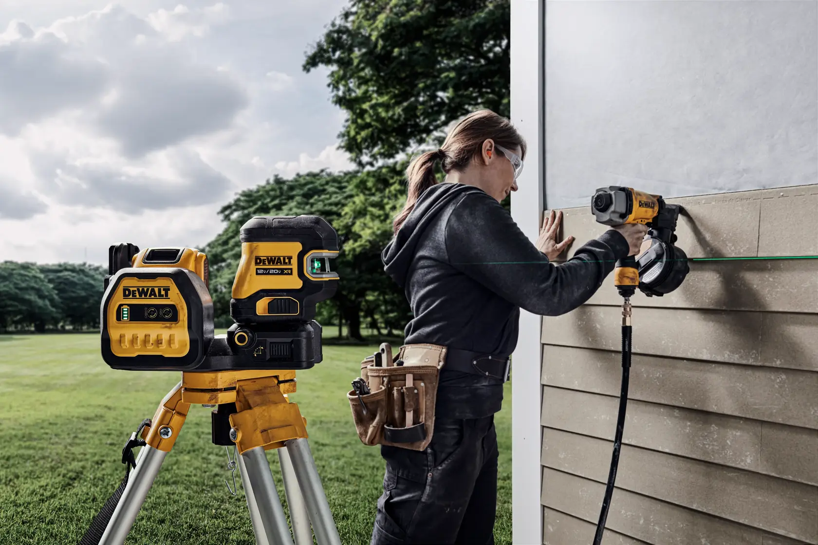 A person with blurred face is using a DeWalt tool to install siding on an exterior wall, with a DeWalt laser level on a tripod in the foreground. The person is wearing work clothes and a tool belt, and is working outdoors near a grassy area and trees.