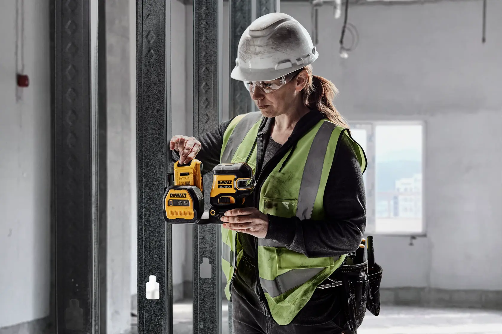 A person wearing a safety vest and hard hat uses a DEWALT laser level in a construction setting. The individual's face is blurred and they are standing indoors next to metal frames.