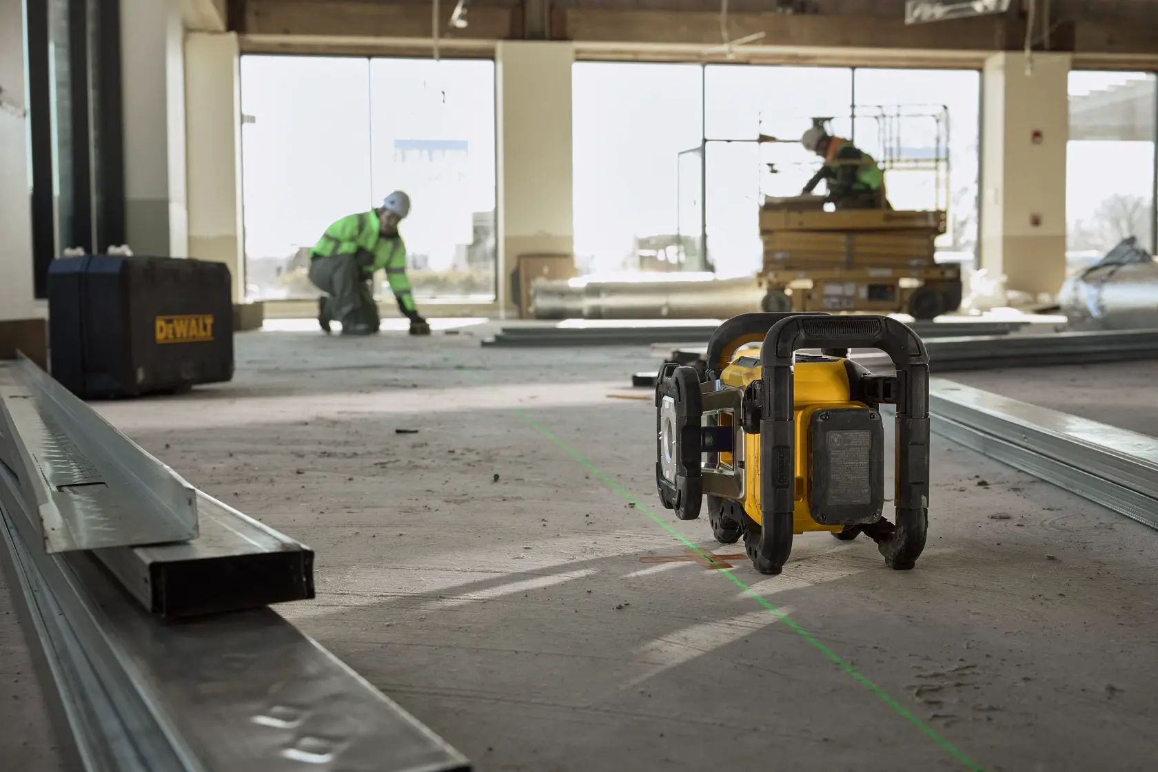 A DEWALT laser level tool is positioned on the floor in a construction site, projecting a green beam line. Metal studs and a DEWALT case are visible, with two workers in safety gear working in the background.
