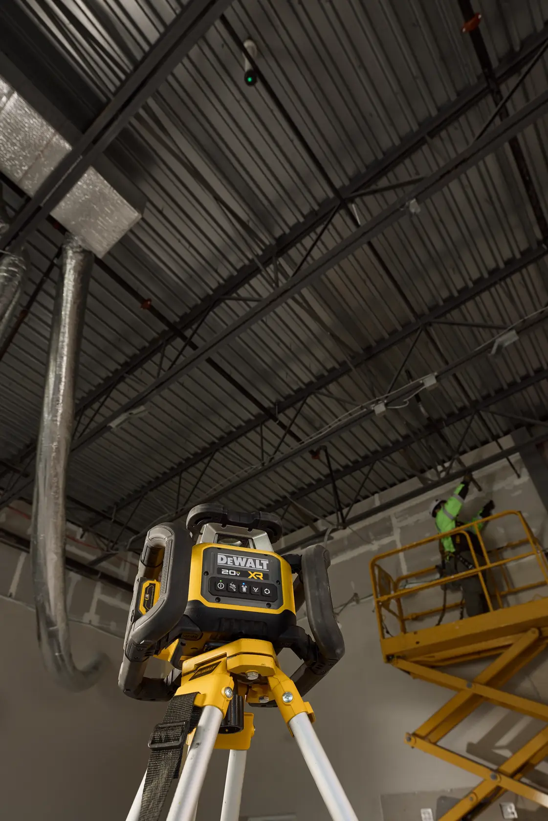 A DEWALT 20V XR laser level is mounted on a tripod in an industrial setting, with a worker on a yellow lift in the background working near the ceiling.