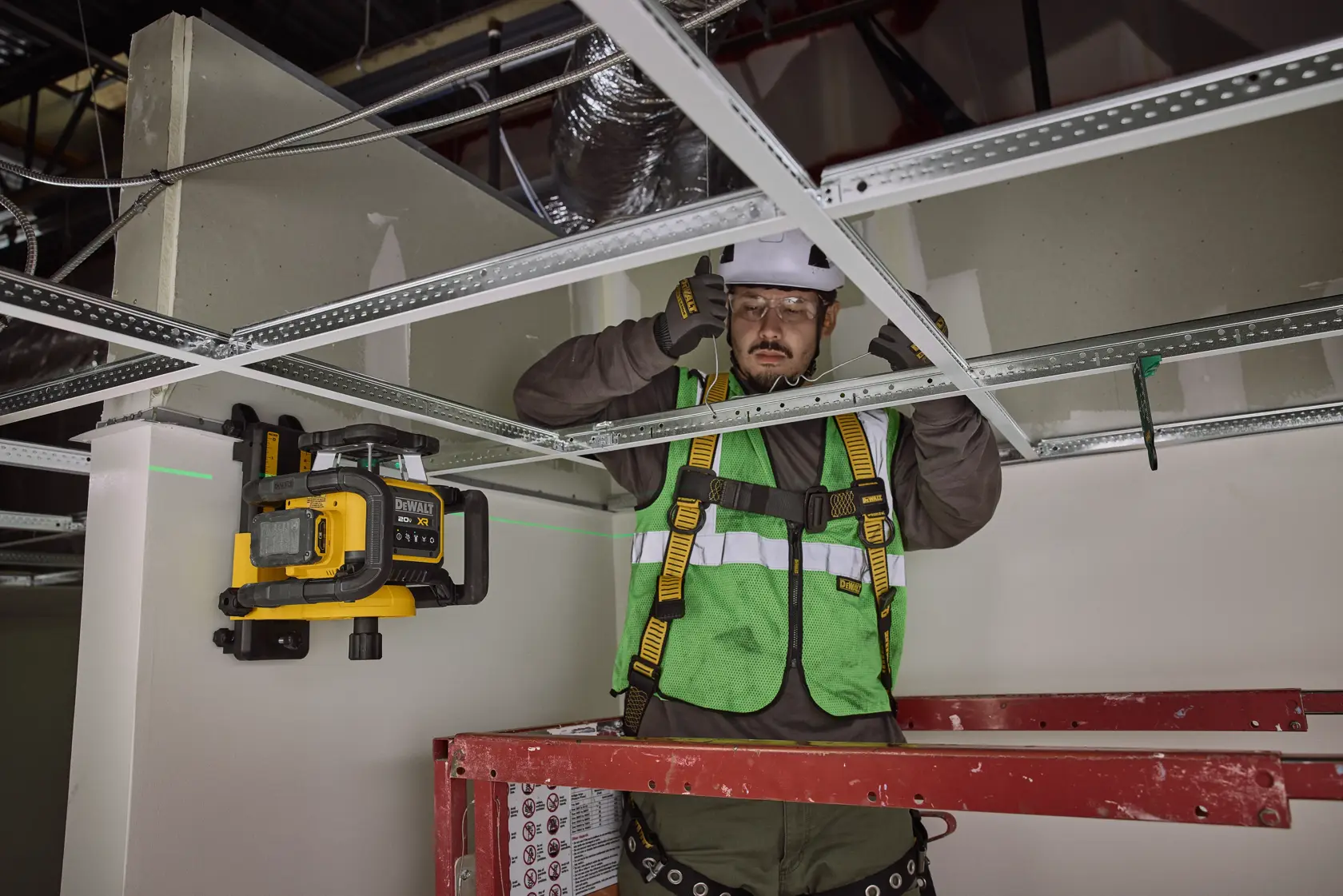A DEWALT laser level device is mounted on a wall, projecting a green laser line for alignment. A construction worker wearing a green safety vest and gloves is installing metal framing overhead, standing on a red ladder in an indoor building under construction.