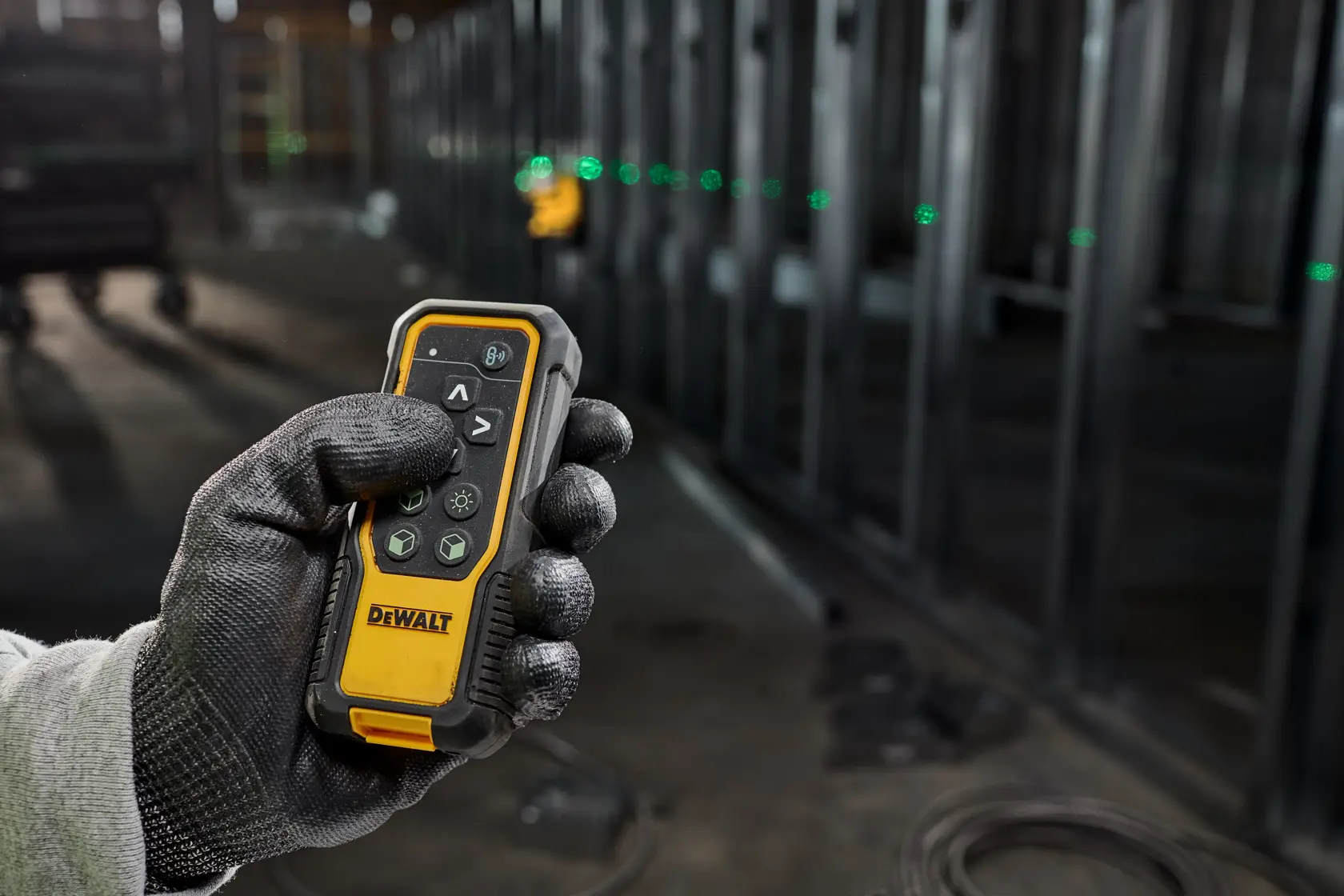 A gloved hand holding a DEWALT laser receiver device, with a DEWALT laser level positioned on a metal frame in the background and visible green laser lines.