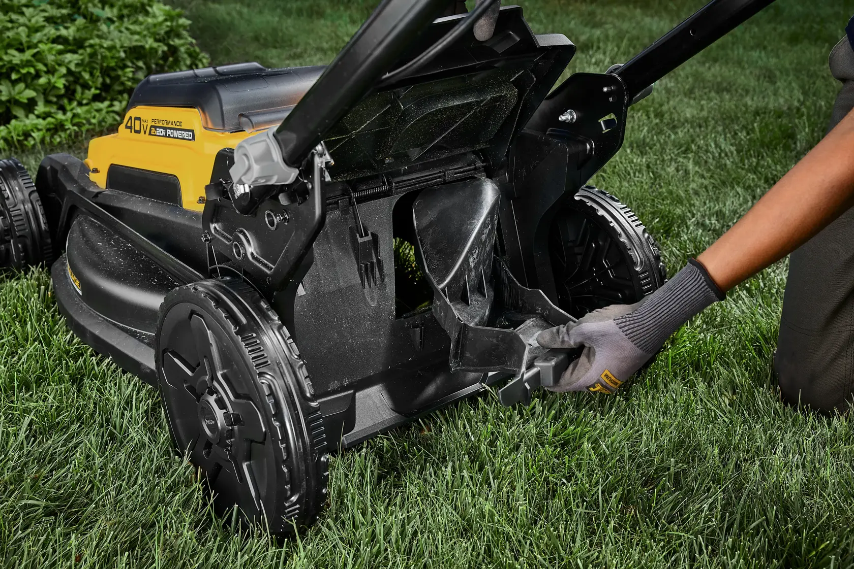 Person adjusts the chute of a yellow DEWALT 40V lawn mower on grass, wearing gloves for safety.