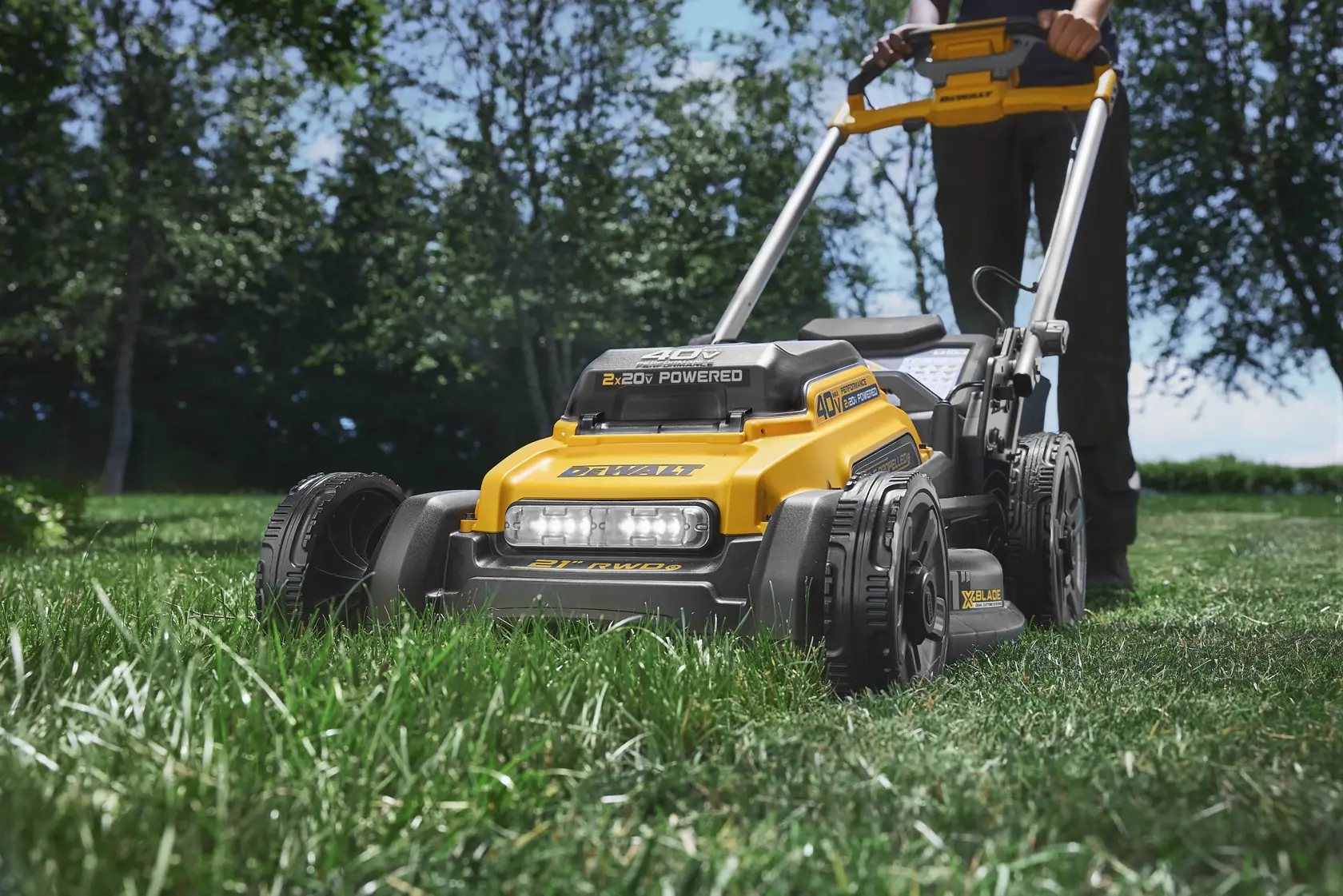 Person using a yellow DEWALT cordless lawn mower on grass in a sunny outdoor setting.