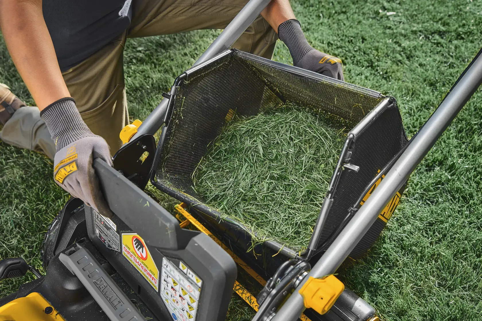 Person empties grass clippings from a DEWALT lawn mower bag onto green lawn, wearing gloves and brown pants.