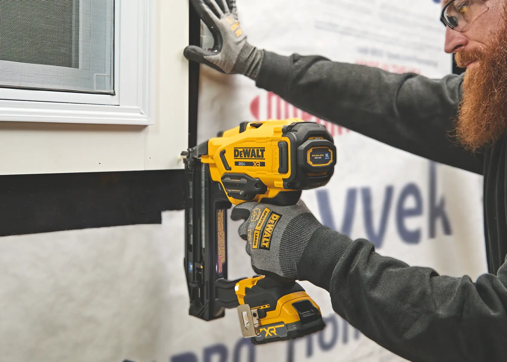Une personne portant des gants de protection utilise un cloueur sans fil DEWALT DCN220 pour installer une moulure autour d’un cadre de fenêtre. L’outil jaune et noir est placé contre le mur lors d’un projet de construction ou de rénovation.