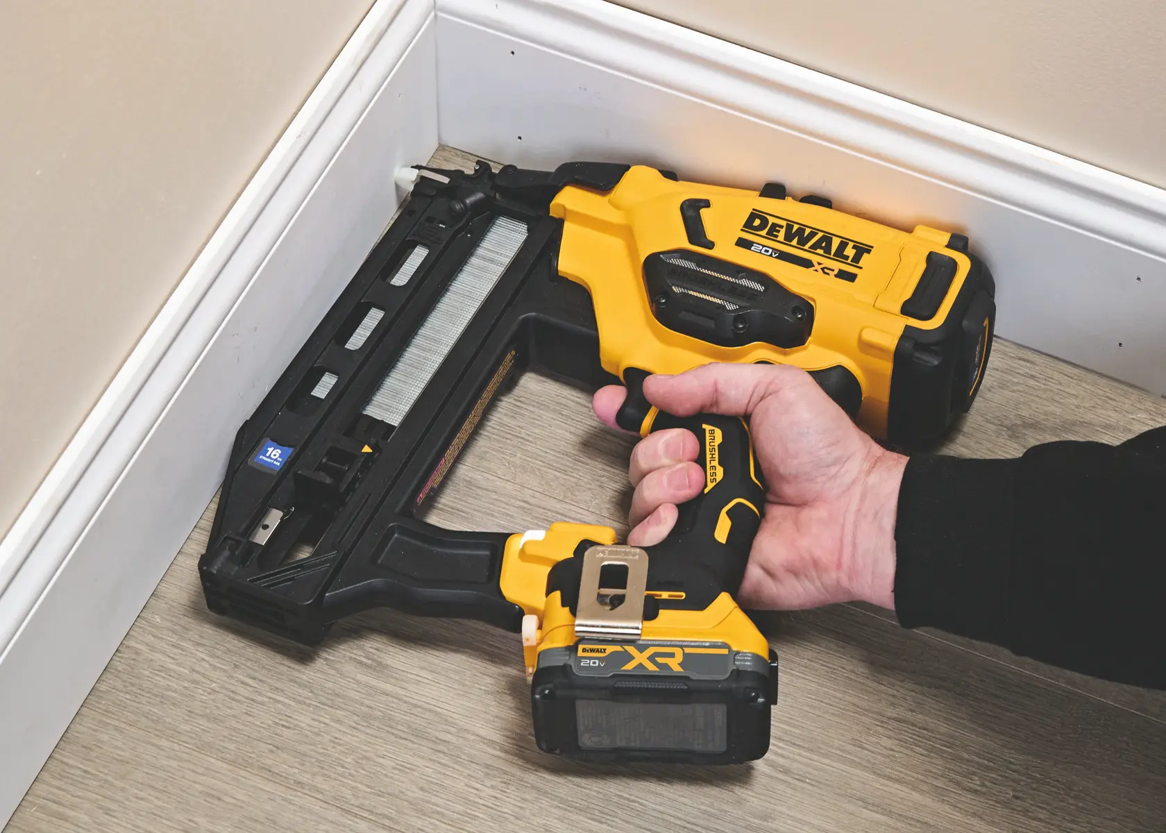 Une main tenant une agrafeuse sans fil DEWALT jaune et noire, utilisée pour fixer une plinthe dans un coin sur un sol de couleur claire.