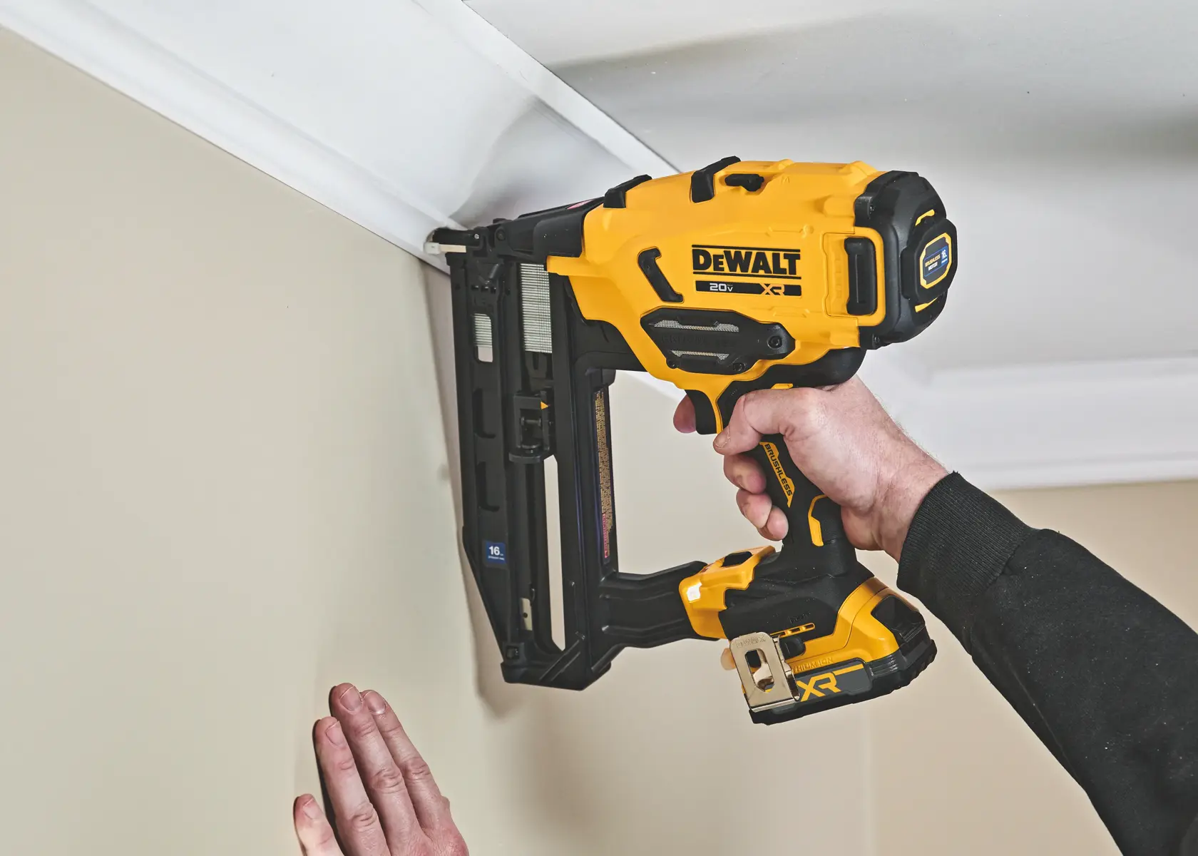 Une personne utilise un pistolet à clous sans fil DEWALT 20V pour fixer une moulure au mur près du plafond.