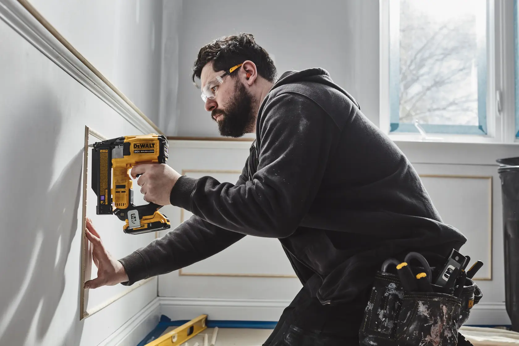 A person using a DEWALT DCN623D1 cordless nailer to install trim on a wall, wearing a tool belt with various tools, inside a room with natural light coming from a window.