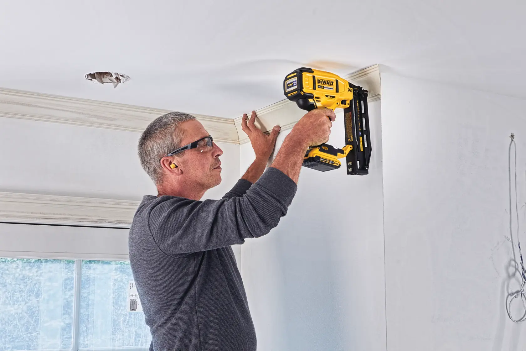 A person is using a DEWALT DCN662D1 nailer to install crown molding on the ceiling of a room. The individual is wearing hearing protection and holding the yellow cordless tool against the molding.