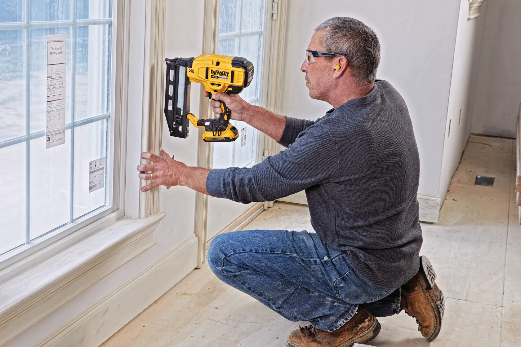 A person kneels while using a DEWALT cordless nail gun (model DCN662D1) to install trim around a window in a residential setting. The person is wearing ear protection and work clothes.