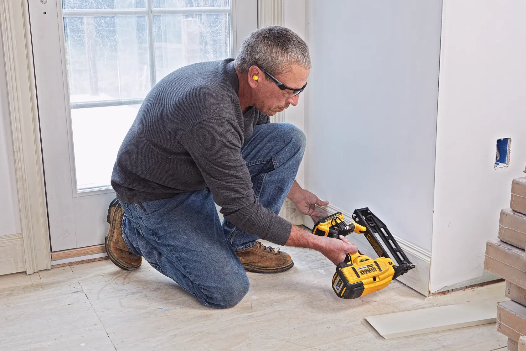 A person kneels on the floor using a DEWALT cordless nail gun, model DCN662D1, to install trim along the base of a wall near a door. The person is wearing ear protection and casual work clothes.