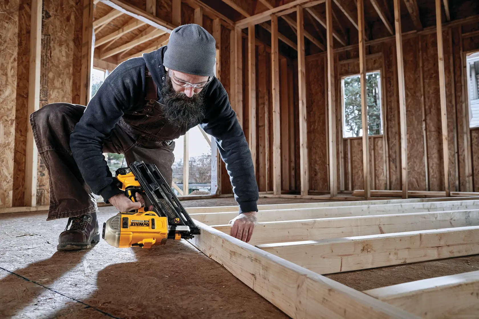 A person uses a DEWALT DCN692M1 cordless nailer to assemble a wooden frame inside a building under construction. The worker is kneeling and wearing a beanie and overalls.