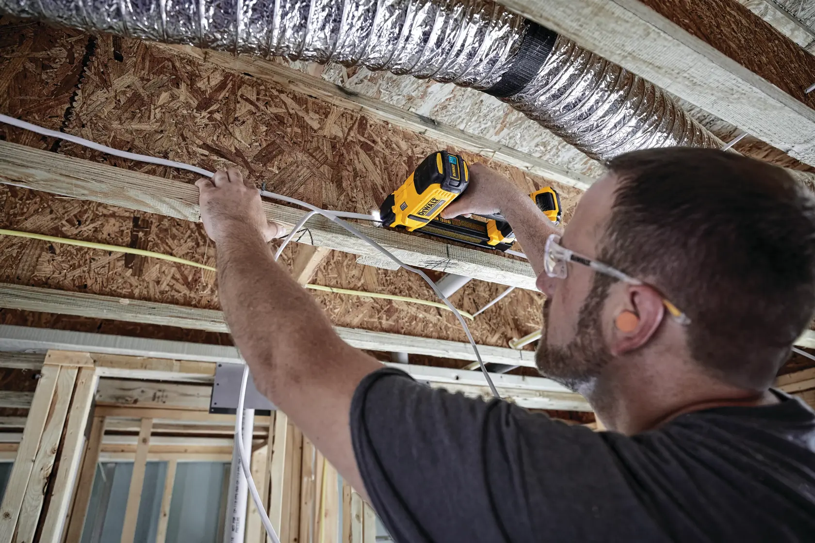 Person using a DEWALT cordless cable stapler to secure electrical wires to wooden beams in a construction setting. The person is holding the stapler against a beam and is wearing safety glasses.