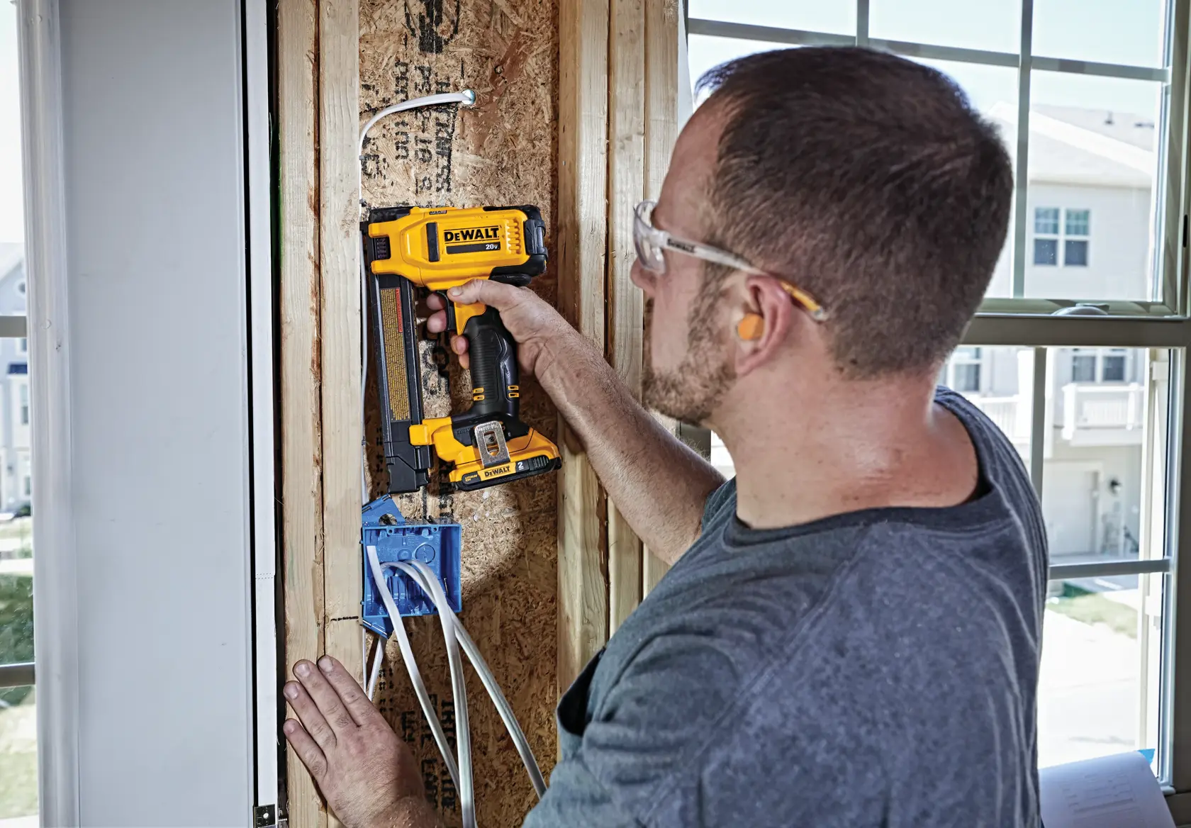A person using a DEWALT DCN701D1 cordless tool to secure electrical wire staples inside a wooden wall frame during construction, with visible wiring and a blue electrical outlet box.