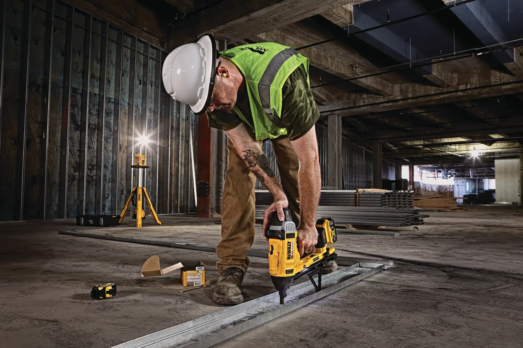 A worker wearing a hard hat and safety vest uses a DEWALT nailer tool (SKU: DCN891B) to fasten metal framing on the floor in a large construction site. Other DEWALT tools and construction materials are visible on the ground.