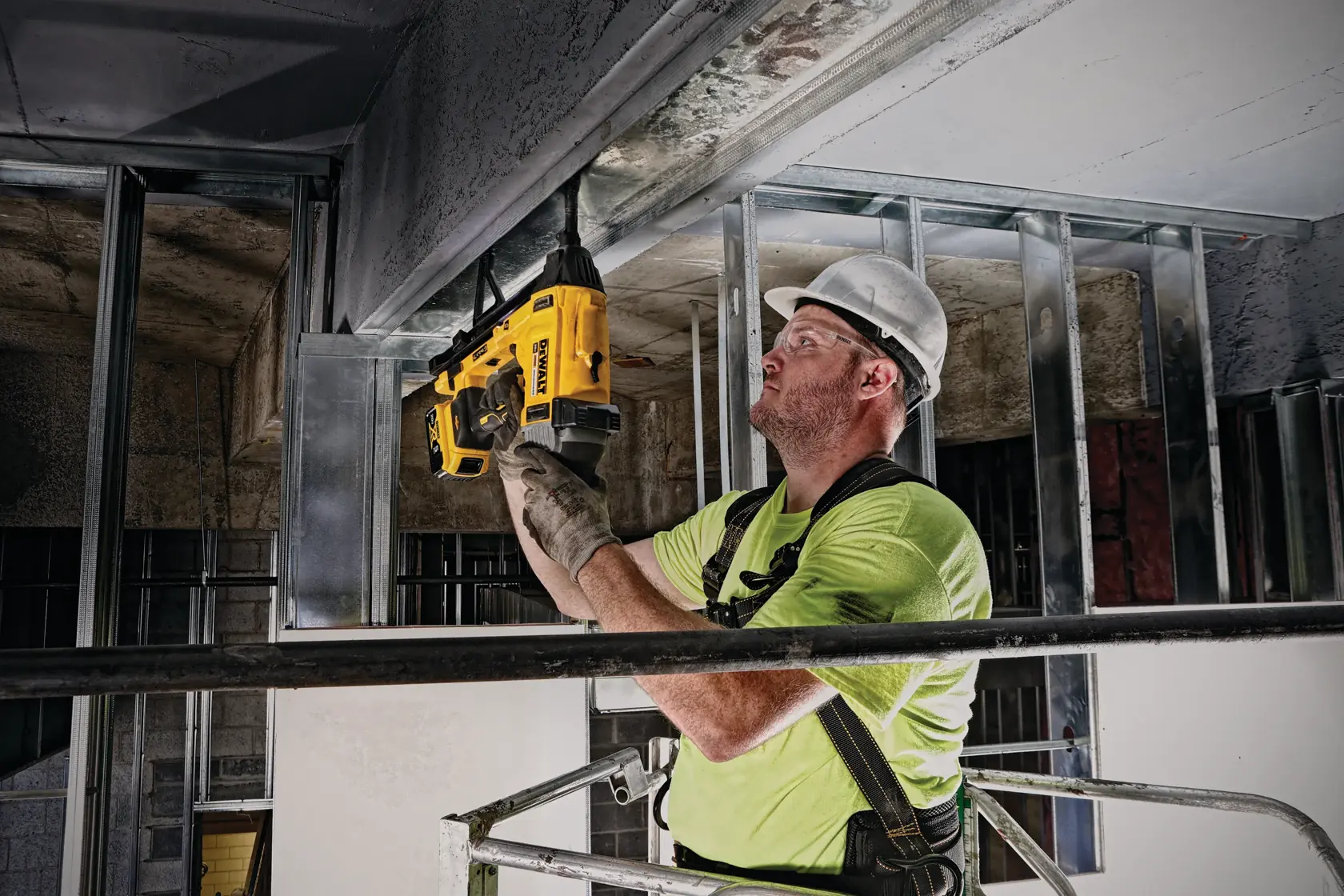A construction worker wearing a hard hat and gloves is using a yellow DEWALT tool, model DCN891B, to secure materials on a ceiling frame at a building site. The worker is standing on a lift and positioned under metal framing.