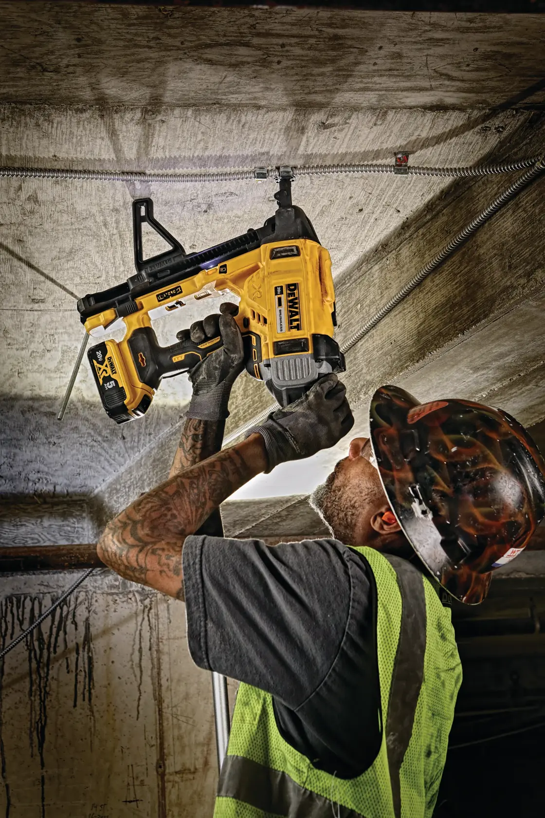 A construction worker wearing a safety vest and hard hat uses a yellow DEWALT tool to fasten components to a concrete ceiling.