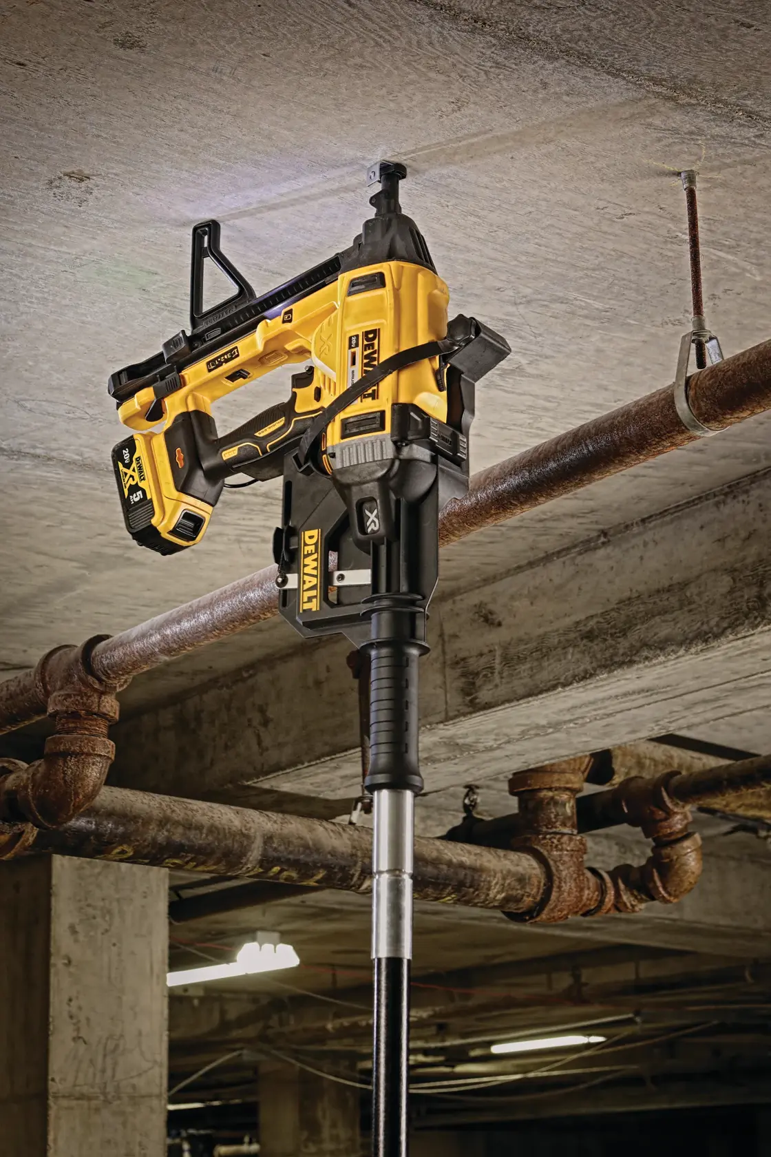 A DEWALT branded cordless tool, model DCN891B, mounted on an extension pole and being used to fasten a pipe clamp to the ceiling of an industrial space.