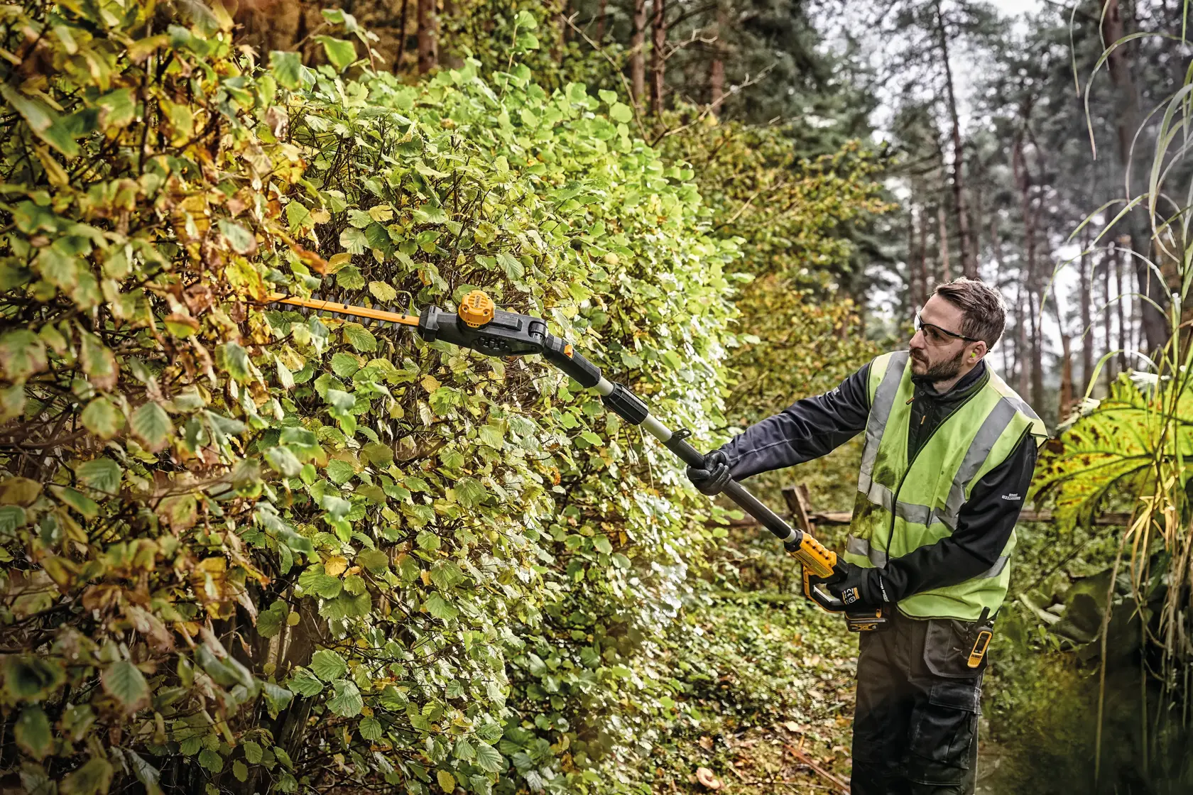 Pole Hedge Trimmer with a battery being used by a person to cut through landscape outgrowth.