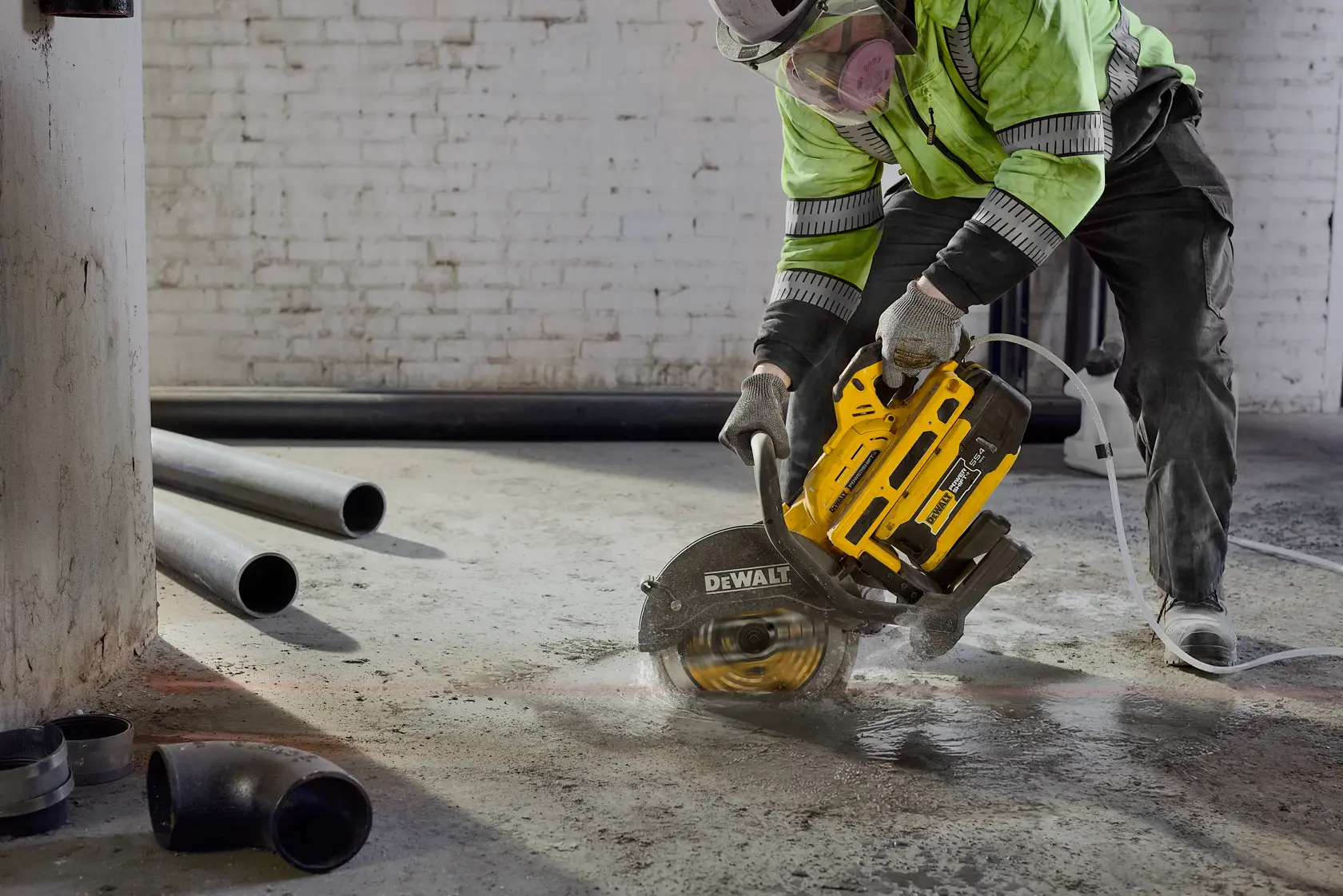 A worker wearing safety gear uses a DEWALT power saw to cut through concrete on a construction site. The saw is creating dust and water is being used to suppress it. Several pipes are lying on the ground nearby.