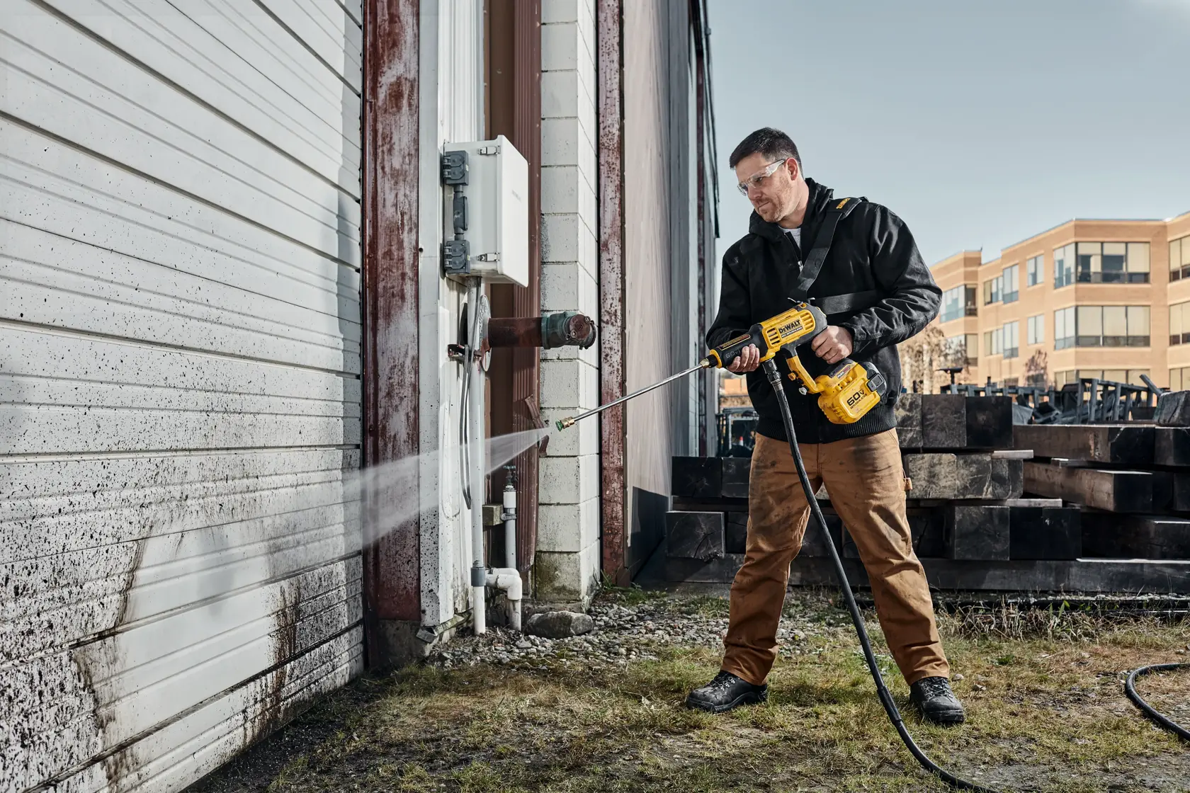 A person is using a DEWALT handheld pressure washer to clean a dirty white garage door outside an industrial building. The person is wearing work clothes and standing on grass near a concrete surface.
