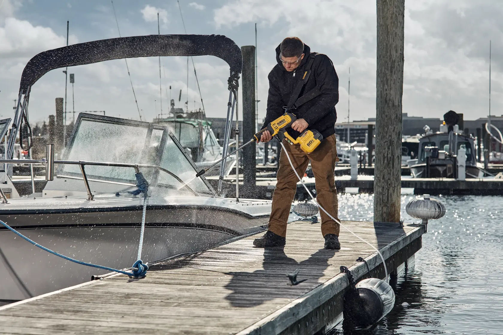 A person is using a yellow DEWALT cordless power washer to clean a boat at a dock. Water is spraying onto the boat, and the person is standing on the wooden dock near the water.