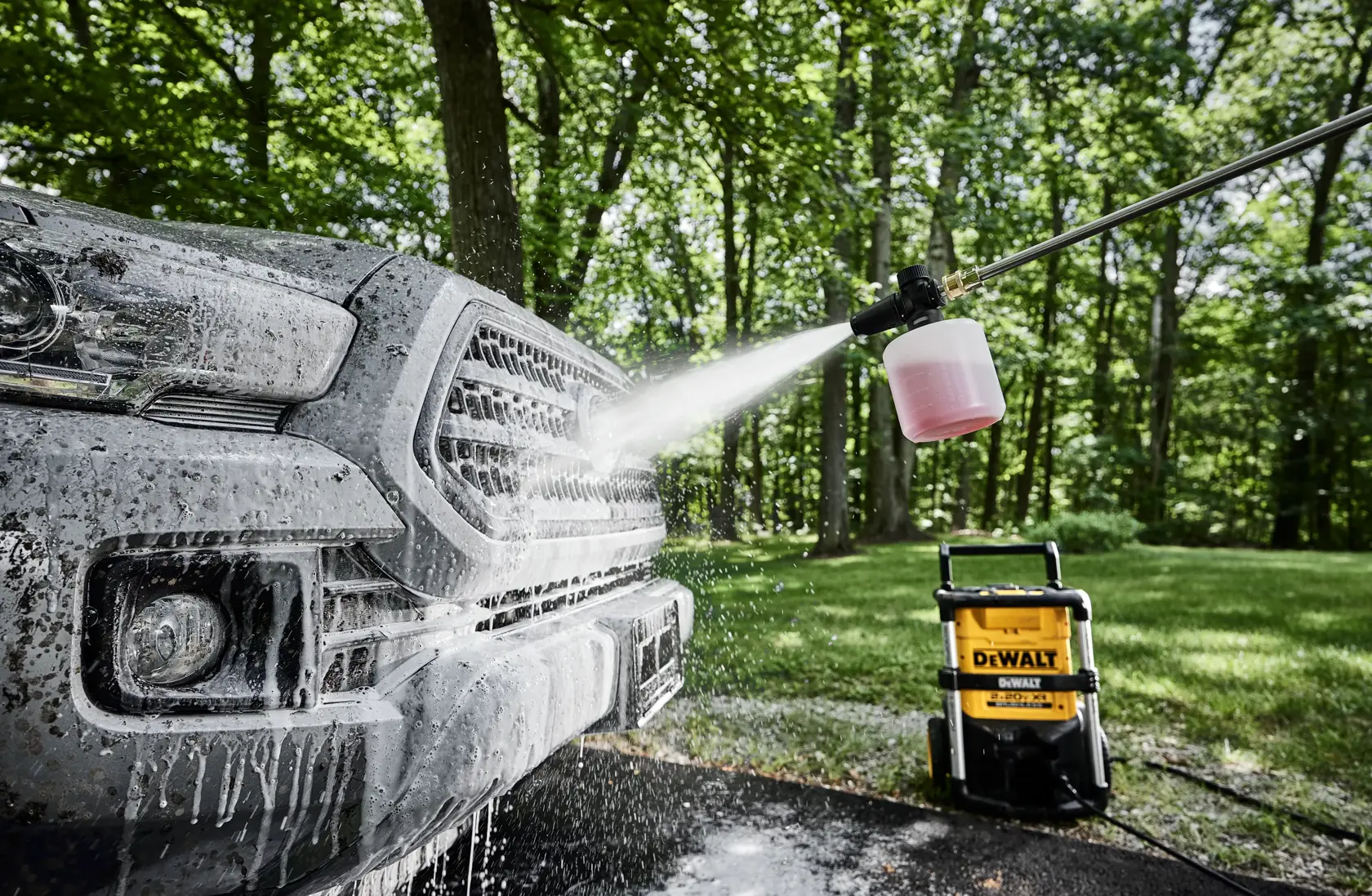 A close-up image of a car being washed with a pressure washer. The pressure washer, branded DEWALT, is spraying foam onto the front grille of the car in an outdoor setting with trees in the background.