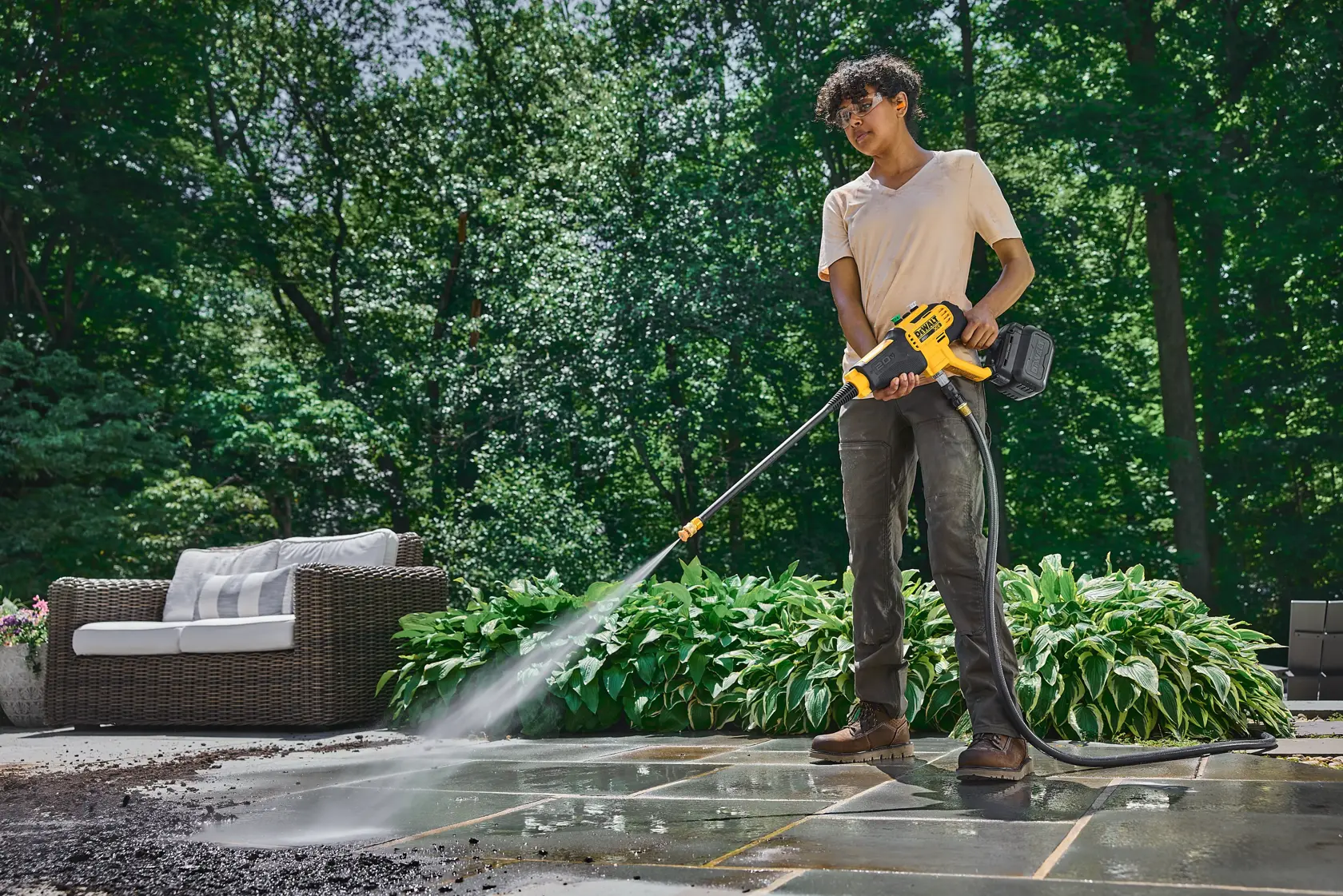 Person cleaning patio with a DEWALT power washer outdoors, surrounded by plants and patio furniture.