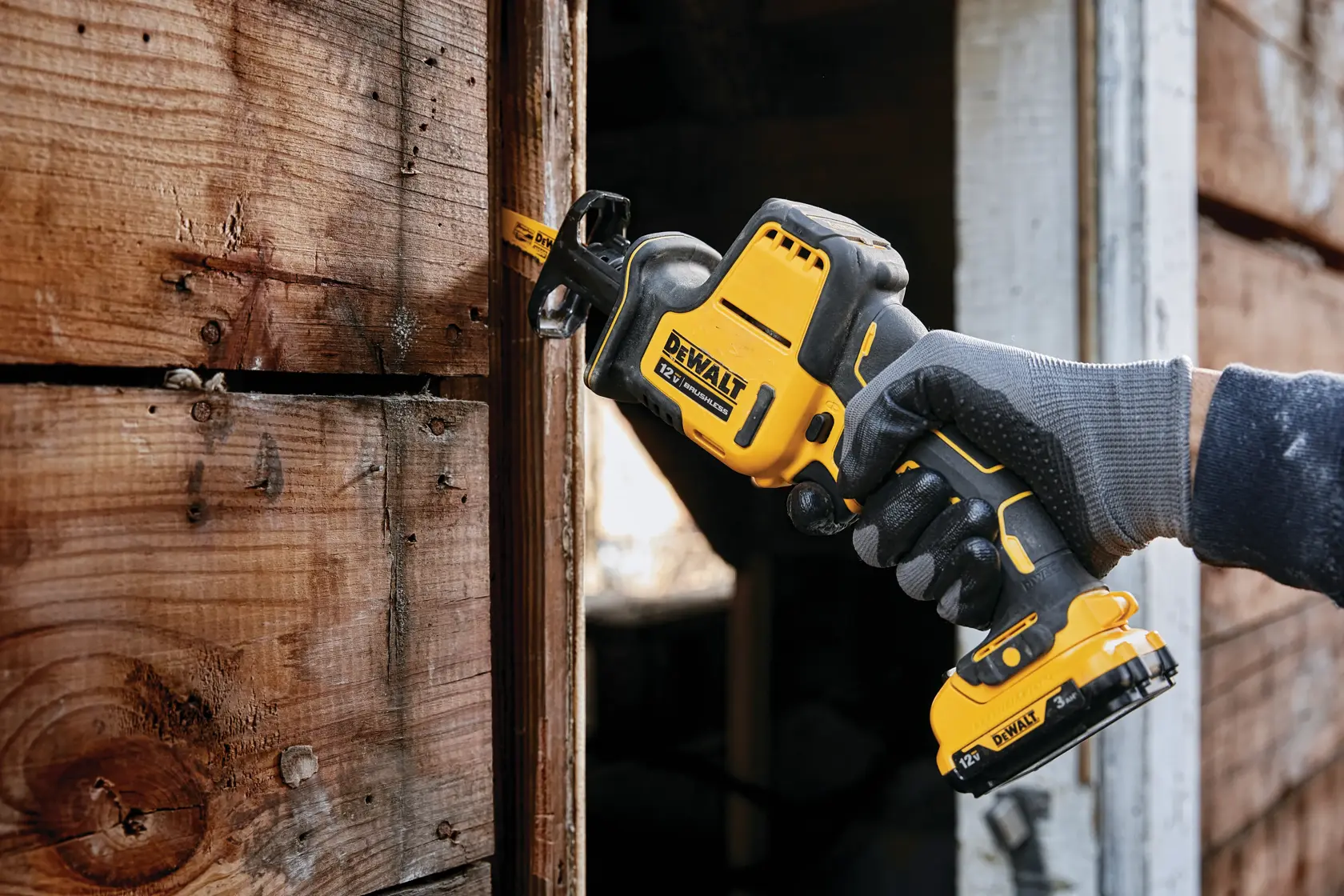 A person wearing gloves uses a DEWALT cordless reciprocating saw to cut through a piece of wood attached to a wooden structure.