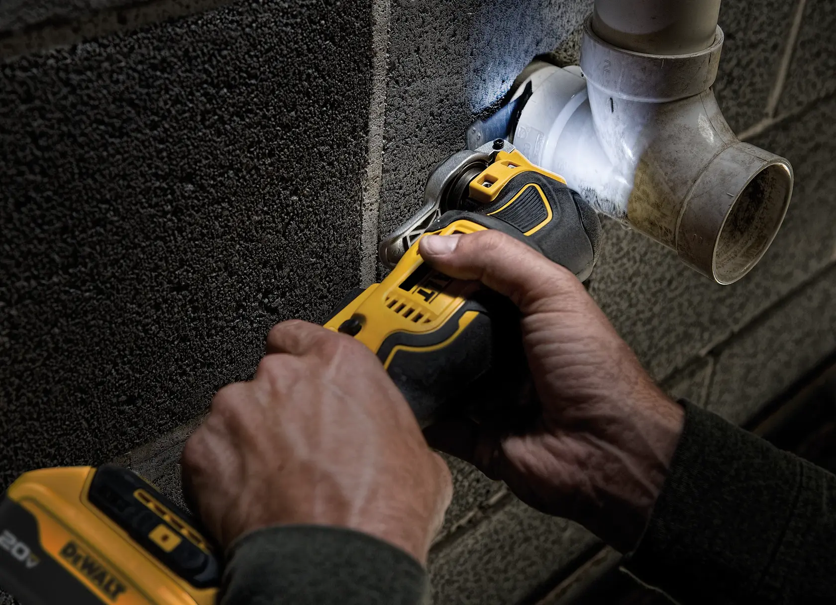 A close-up of hands using a DEWALT DCS356P1 oscillating multi-tool to cut a pipe attached to a concrete wall. The tool is yellow and black and is illuminated by a built-in LED light.