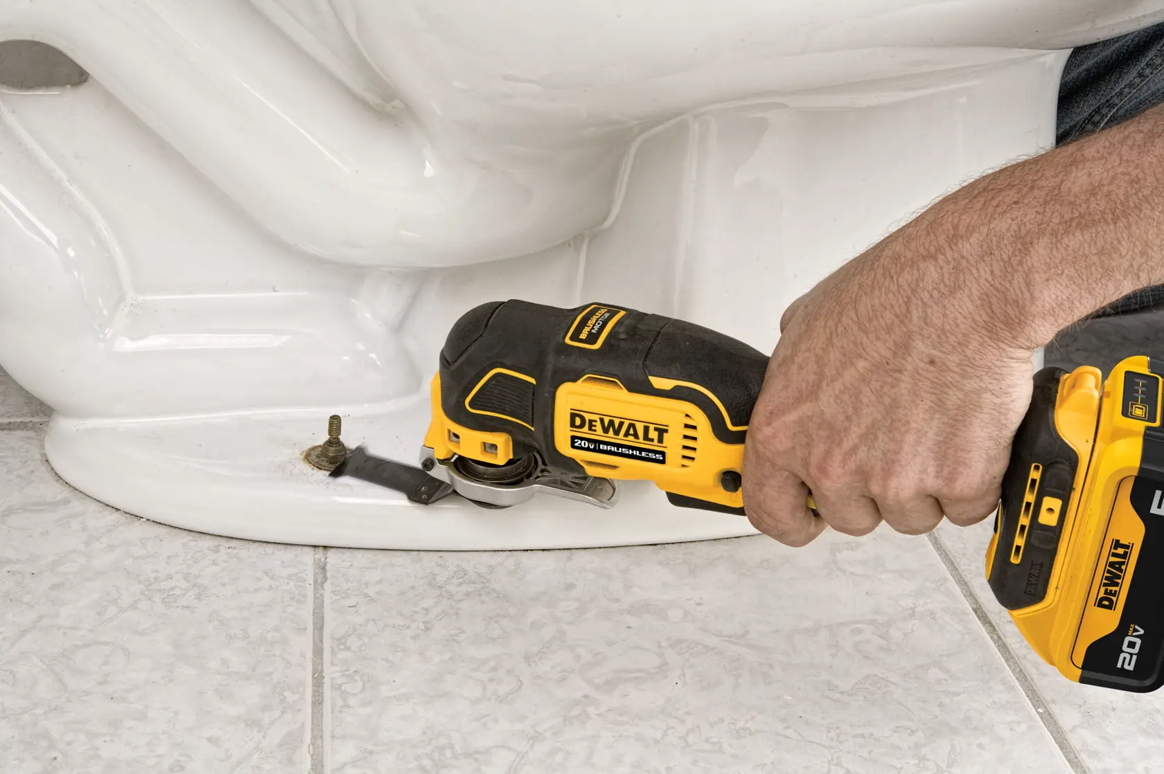 A person uses a DEWALT 20V oscillating multi-tool to cut around a bolt at the base of a ceramic toilet on a tiled floor.