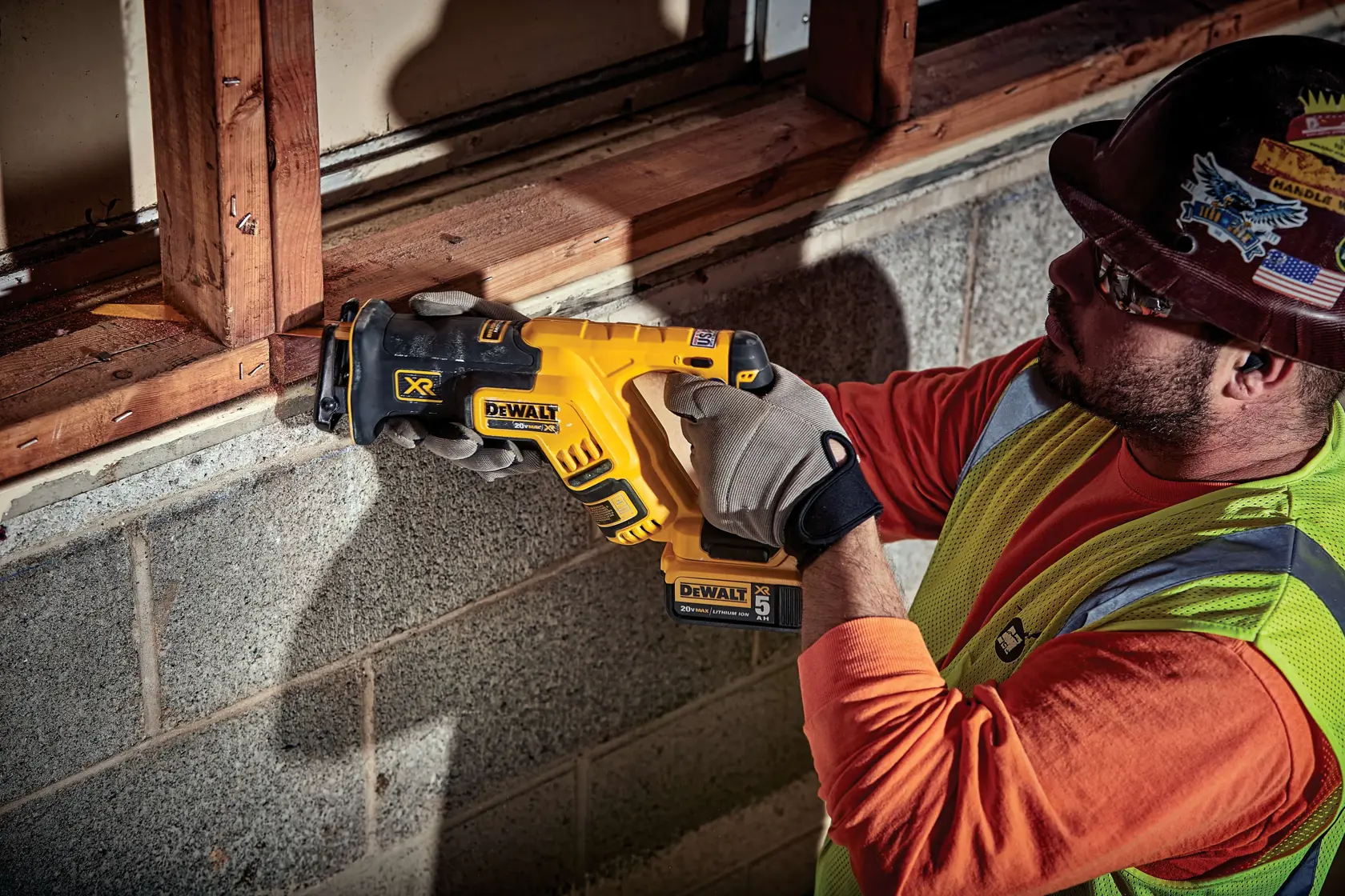 A person wearing work gloves and a safety vest is using a DEWALT DCS367B cordless reciprocating saw to cut a piece of wood at a construction site. The person’s face is obscured for privacy.