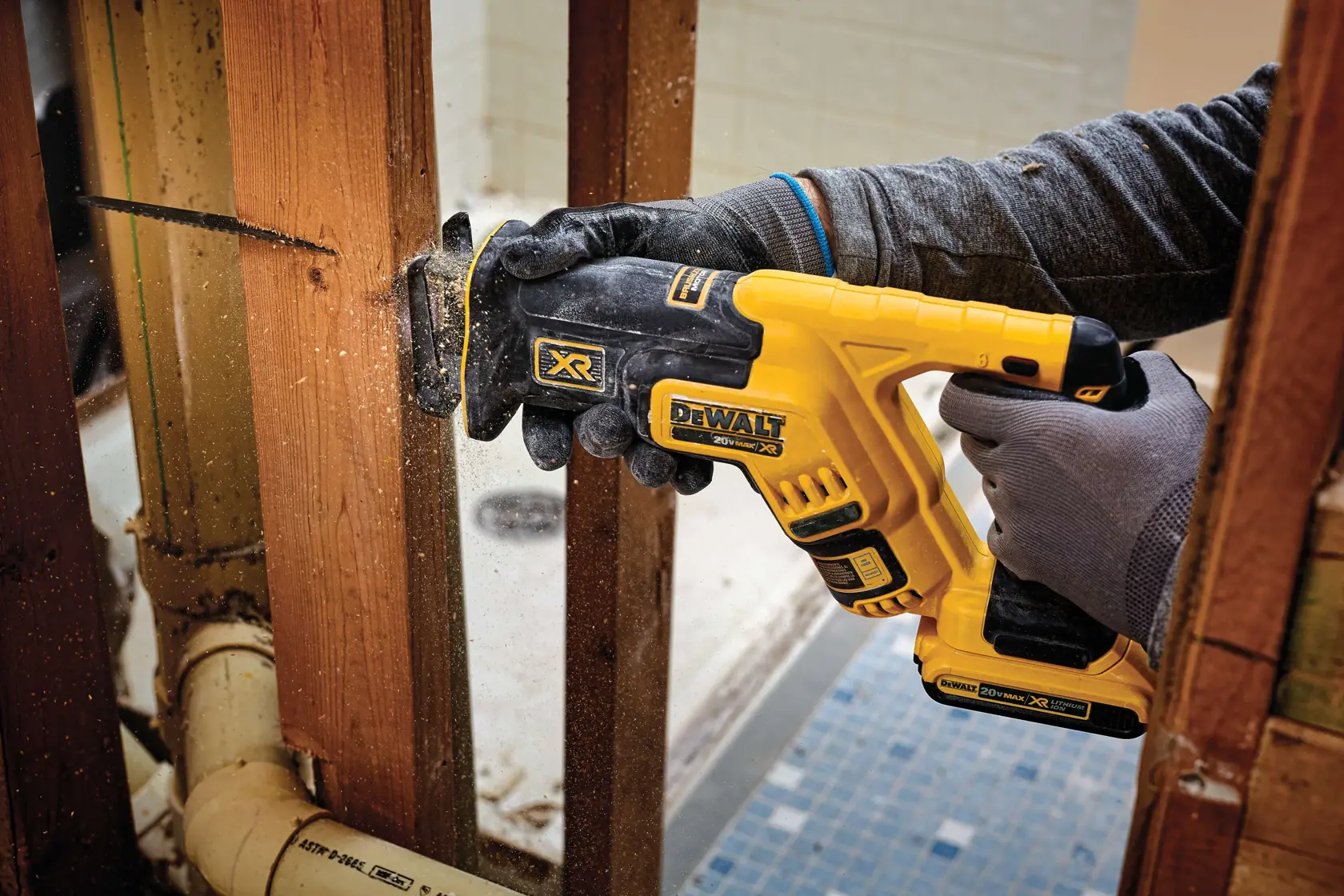 A person wearing gloves uses a yellow and black DEWALT cordless reciprocating saw to cut through a wooden beam inside a building under construction.