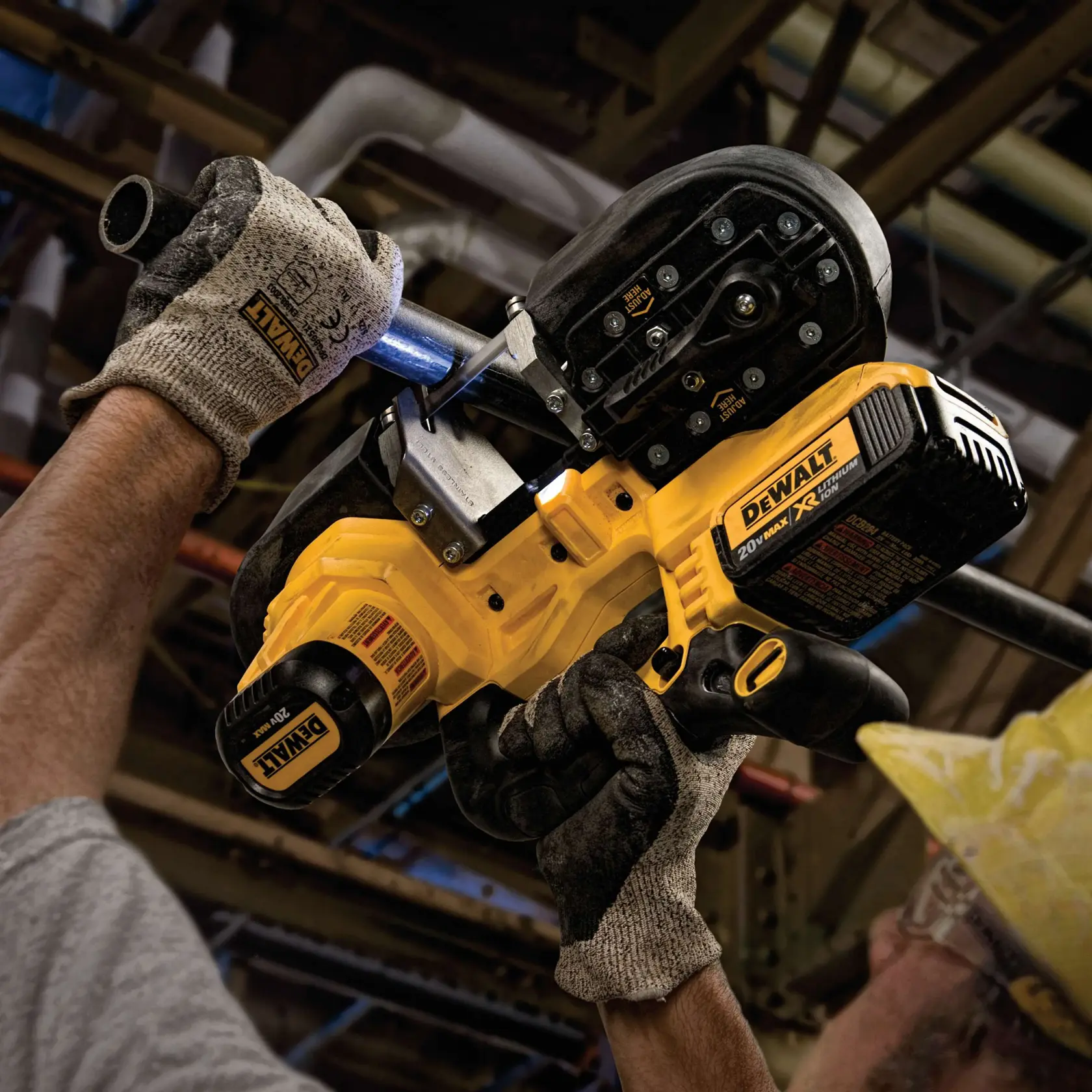 A person wearing work gloves uses a DEWALT cordless portable band saw to cut through a metal pipe in an industrial setting. The tool is yellow and black, labeled with 20V MAX XR Lithium Ion.