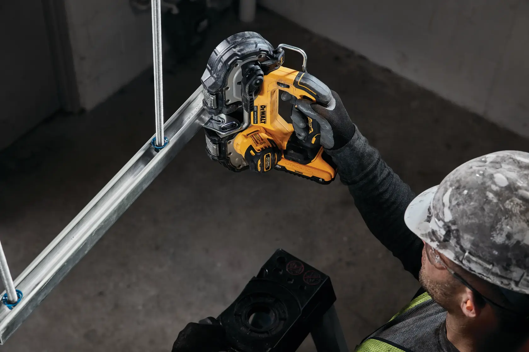 A person wearing a construction helmet and gloves uses a yellow DEWALT portable band saw to cut a metal pipe indoors.