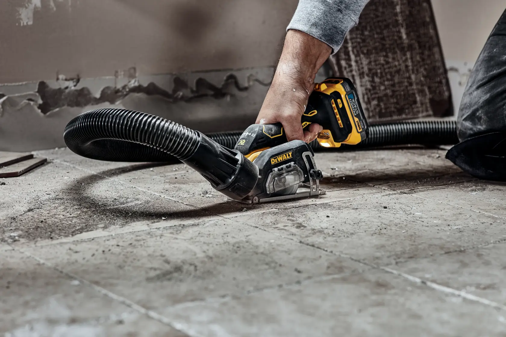 A person using a DEWALT cordless saw with dust extraction attachment to cut or grind tiles on a floor.