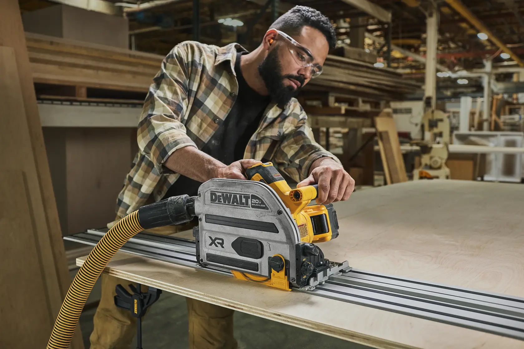 Une personne utilise une scie à rail DEWALT DCS525 XR avec un tuyau de raccordement pour découper une grande planche de bois dans un atelier. Le visage de la personne est masqué et elle porte une chemise à carreaux.