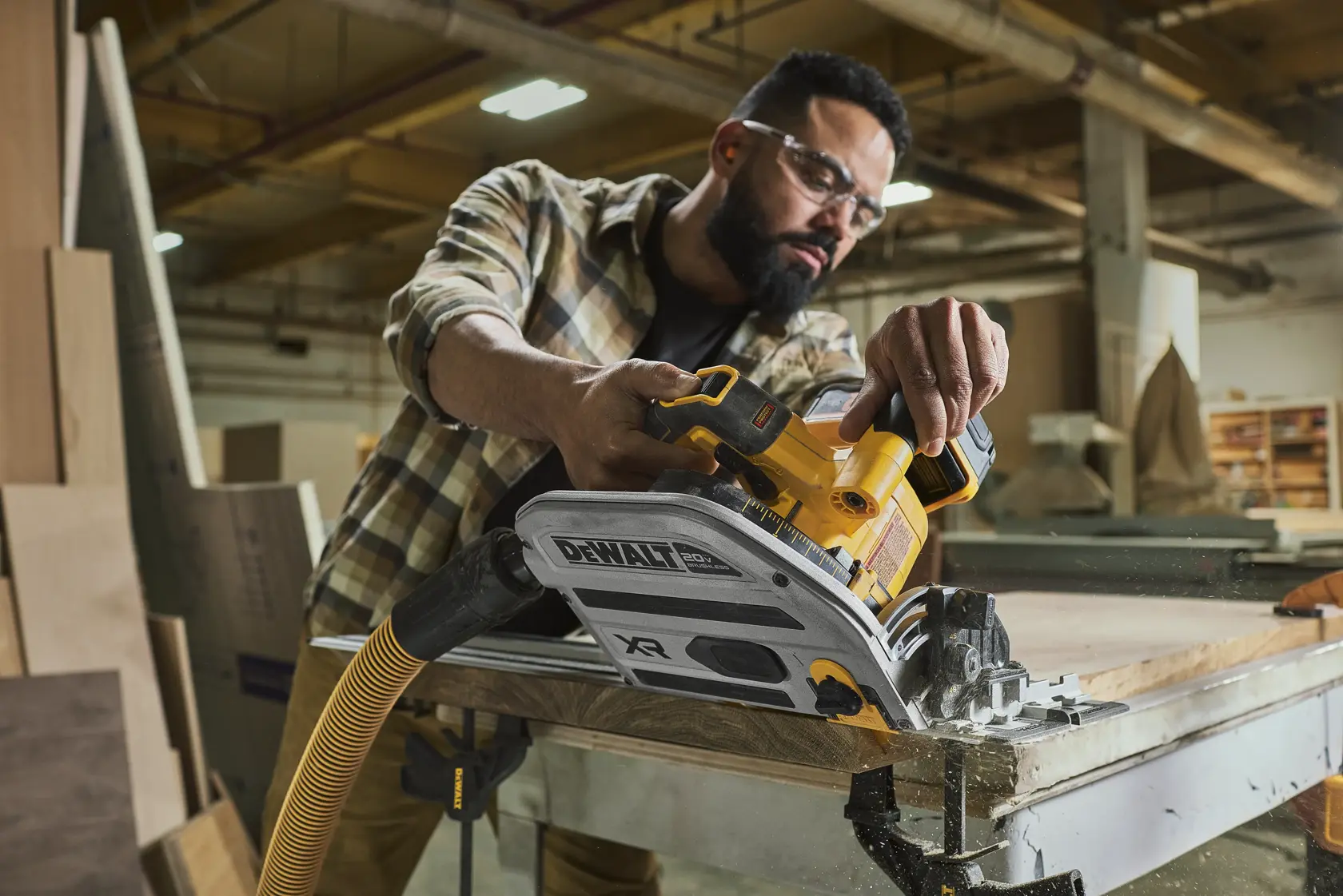 Une personne utilise une scie circulaire sans fil DEWALT DCS525 pour couper une planche de bois dans un atelier. La scie est reliée à un tuyau d’aspiration de poussière, et l’espace de travail contient diverses pièces de bois. Le visage de la personne est flouté pour préserver sa confidentialité.
