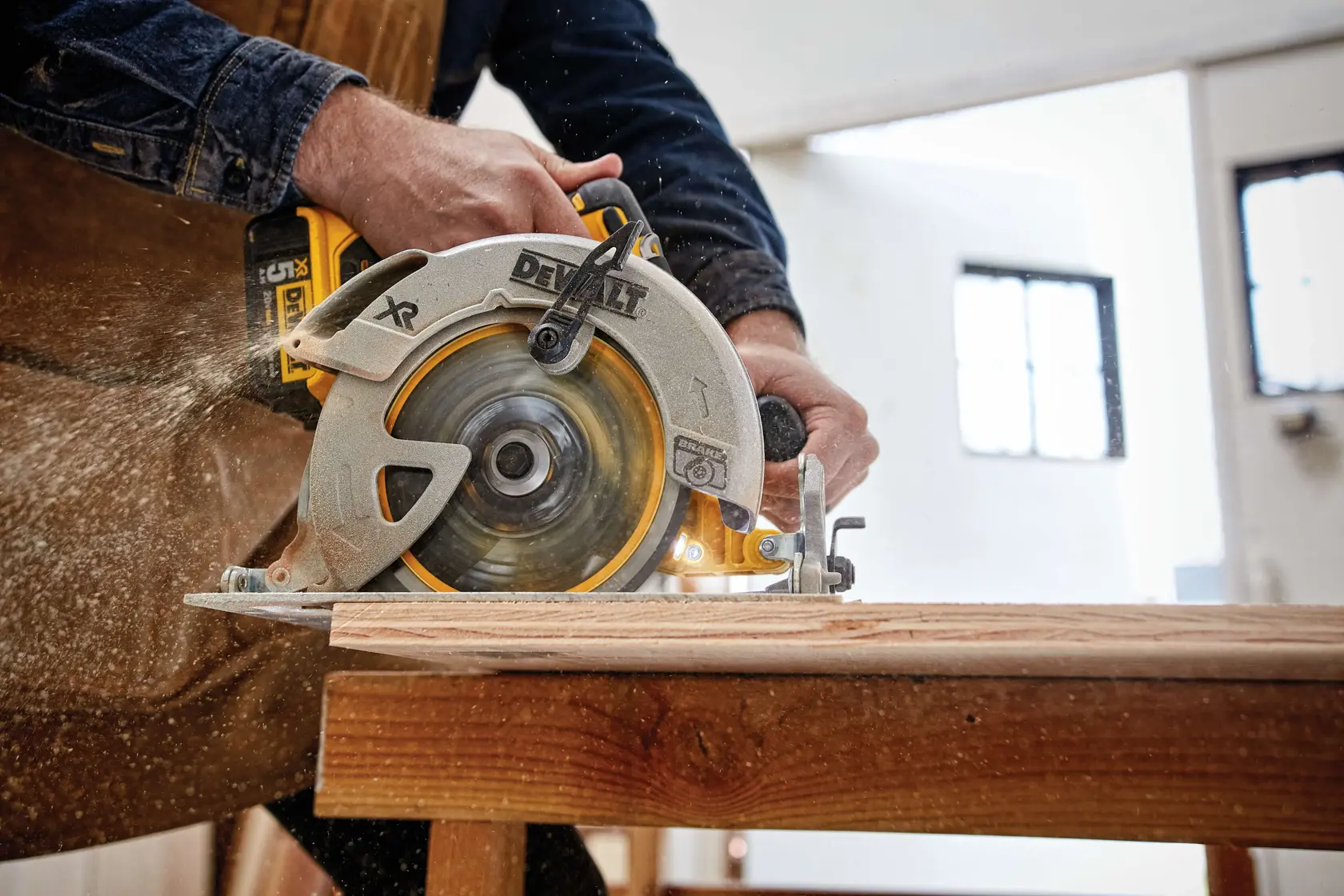 A person is using a DEWALT circular saw to cut a piece of wood on a workbench, with sawdust flying, in a workshop setting.