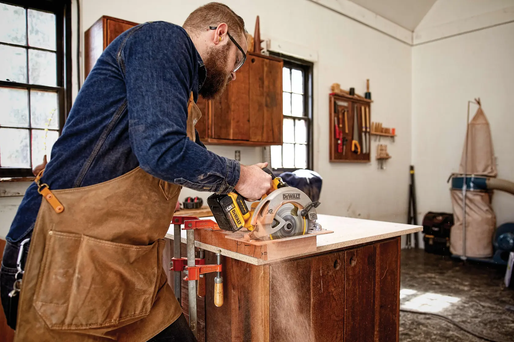 A person wearing a brown apron is using a DEWALT cordless circular saw (model DCS570P1) to cut a piece of wood in a workshop. The workspace includes wooden cabinets, clamps, tools hanging on the wall, and a dust collection system, with sunlight coming in through the windows.