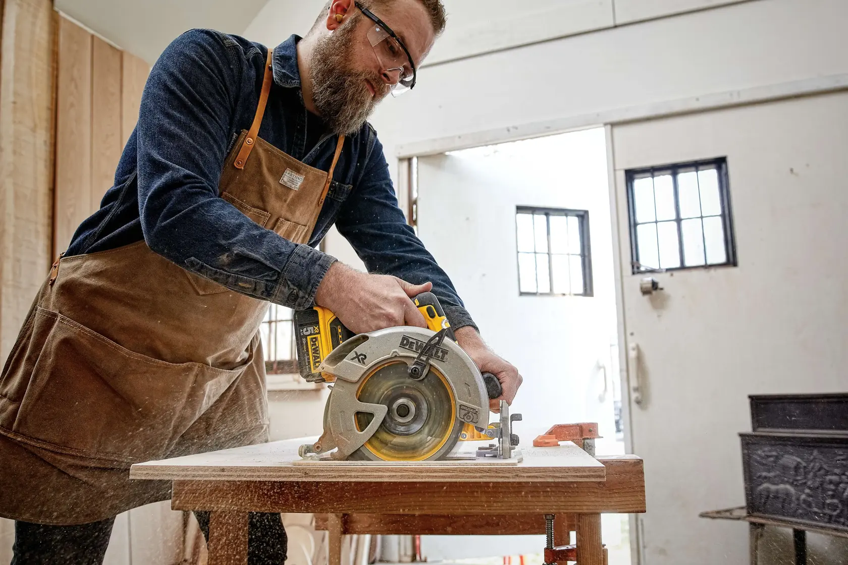 A person wearing a brown apron operates a DEWALT circular saw to cut a wooden board on a workbench in a workshop setting. The person's face is blurred for privacy.