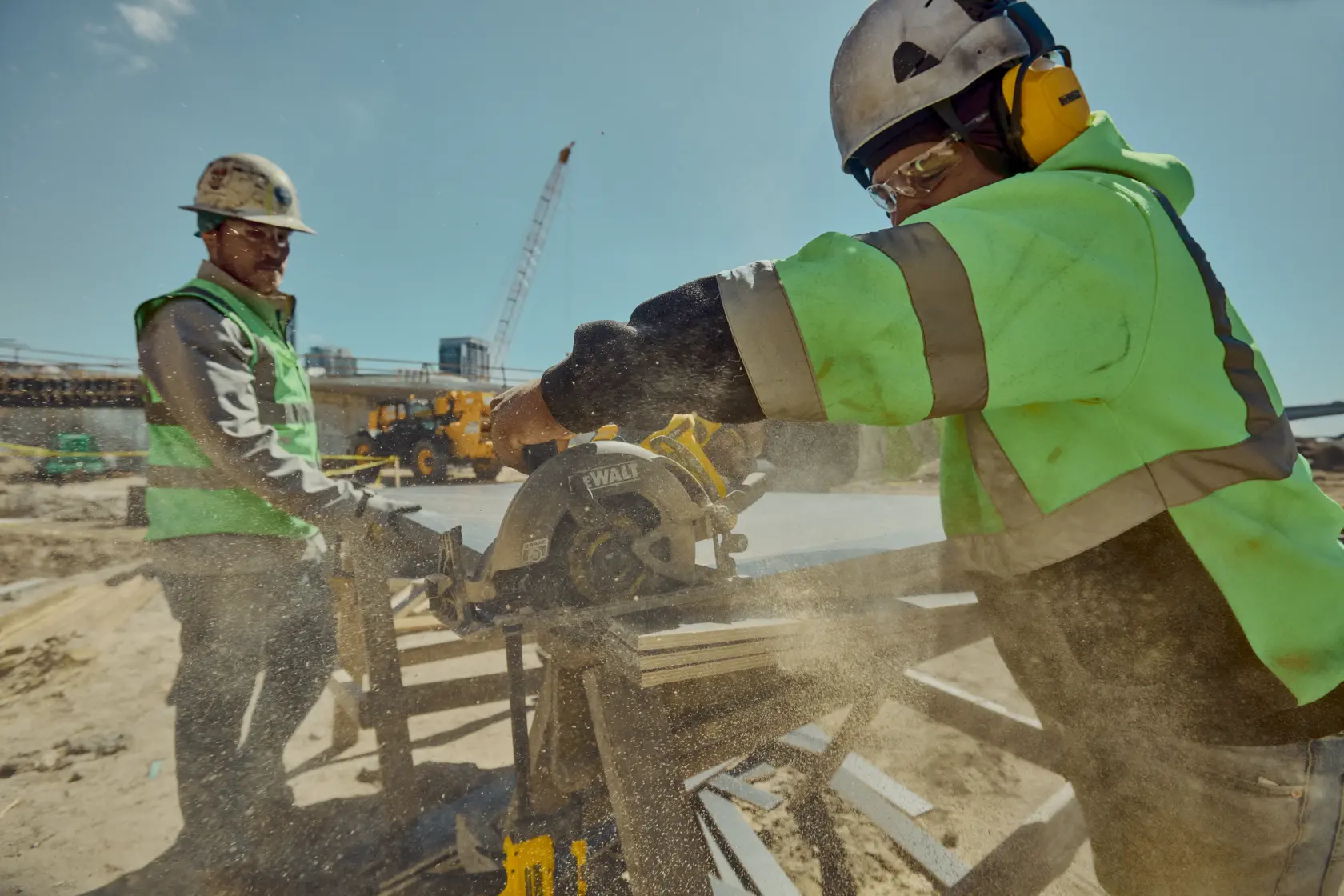 Two construction workers wearing safety gear are using a DEWALT circular saw to cut wood at a busy outdoor construction site. Sawdust is visible in the air around them.