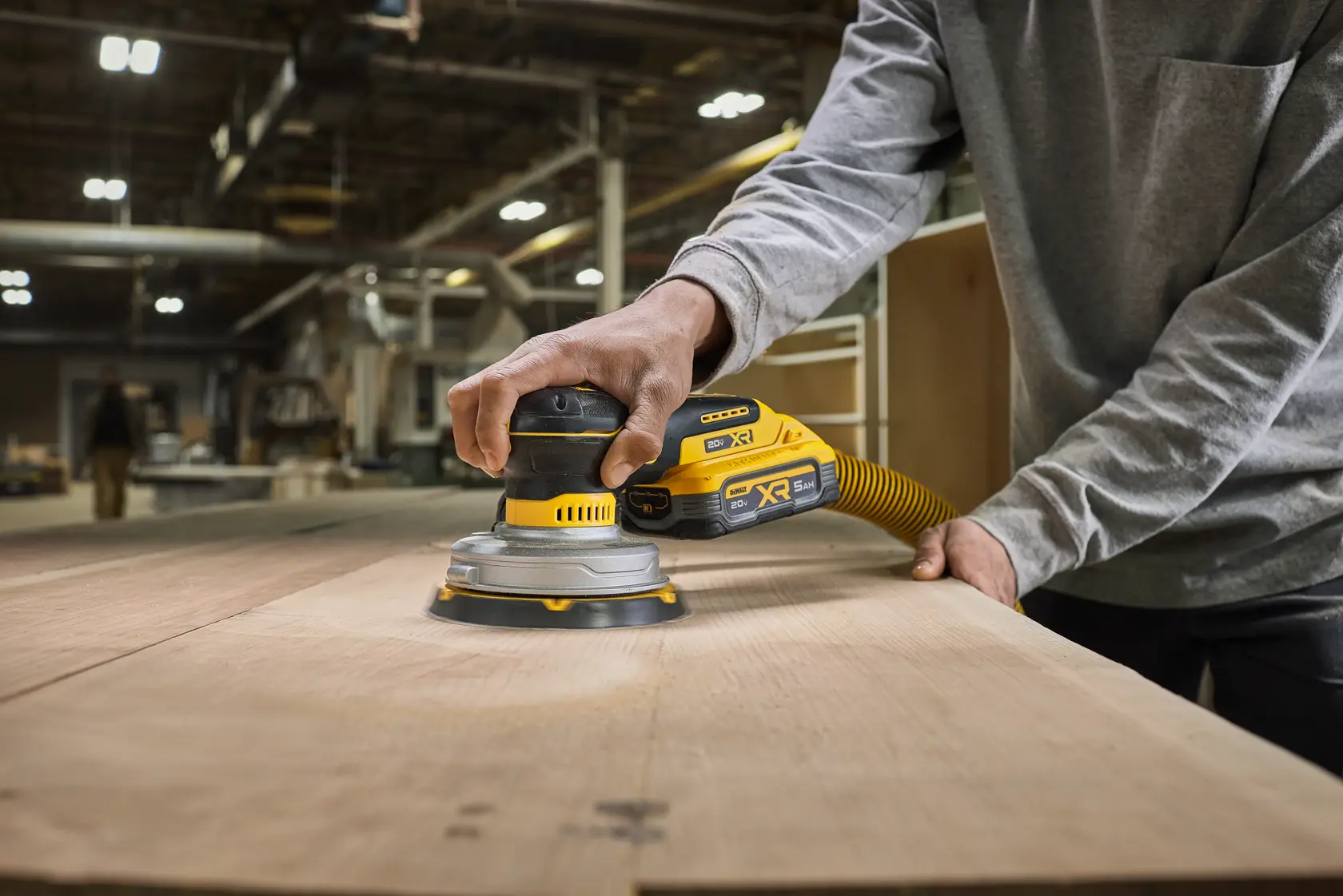 A person using a DEWALT DCW230B cordless sander with a dust extraction hose to smooth a large wooden surface in a workshop environment.