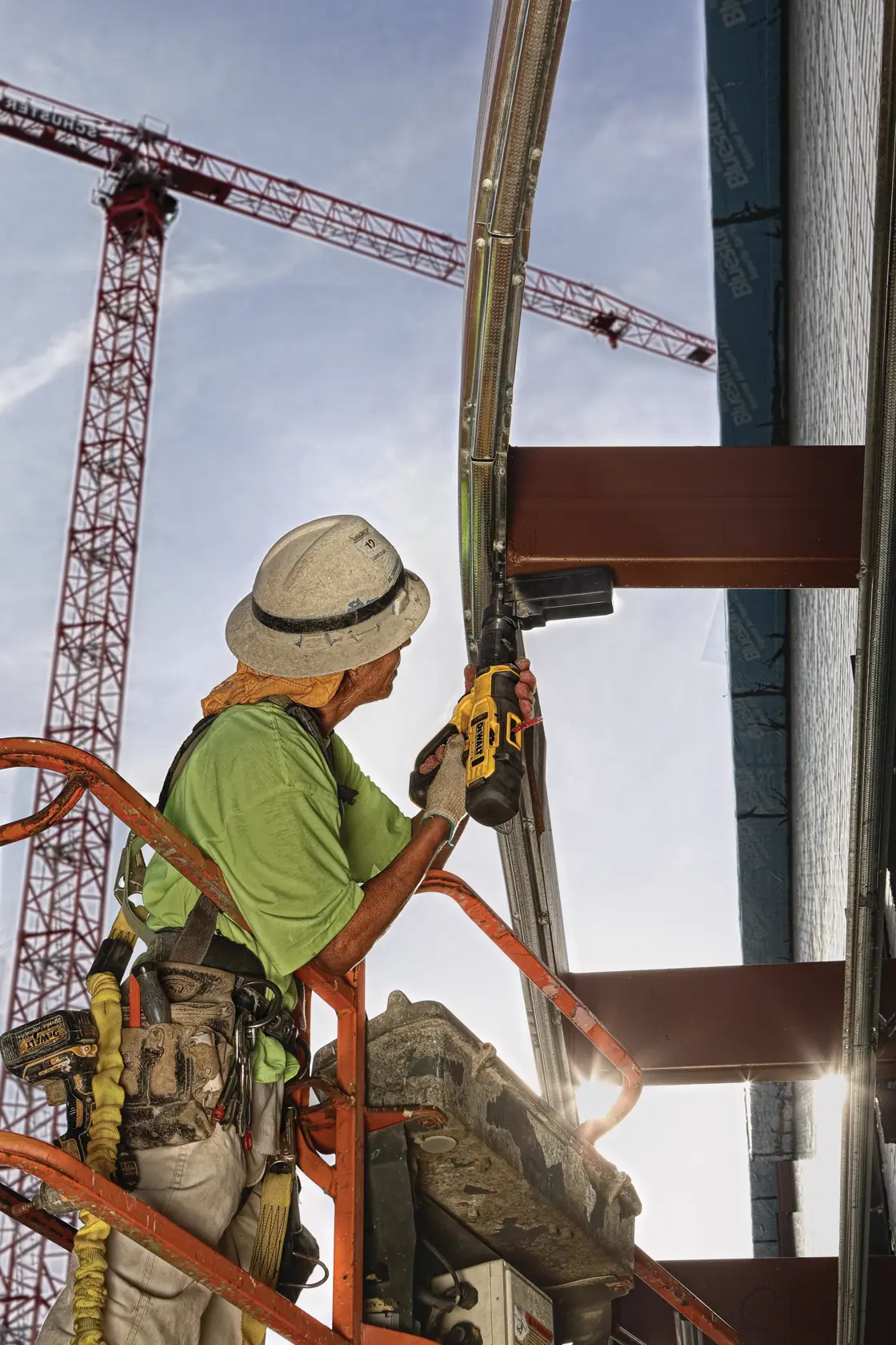 A construction worker in safety gear uses a DEWALT tool to fasten metal on a building structure, standing on an elevated platform with cranes and steel beams visible in the background.