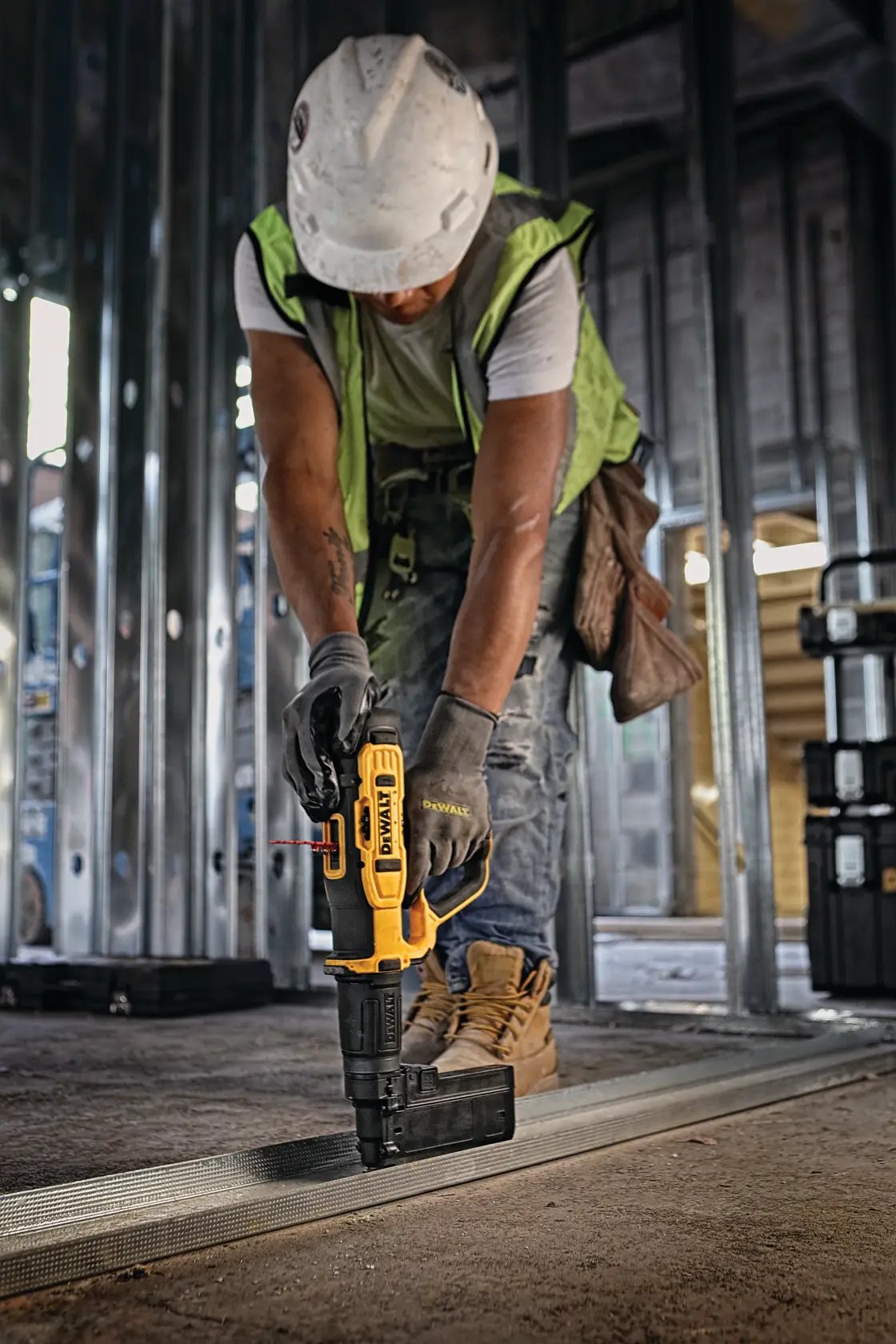 A construction worker wearing a hard hat, safety vest, gloves, and boots is using a yellow DEWALT tool to fasten metal framing on a construction site. The worker is focused on the task and surrounded by unfinished metal walls.